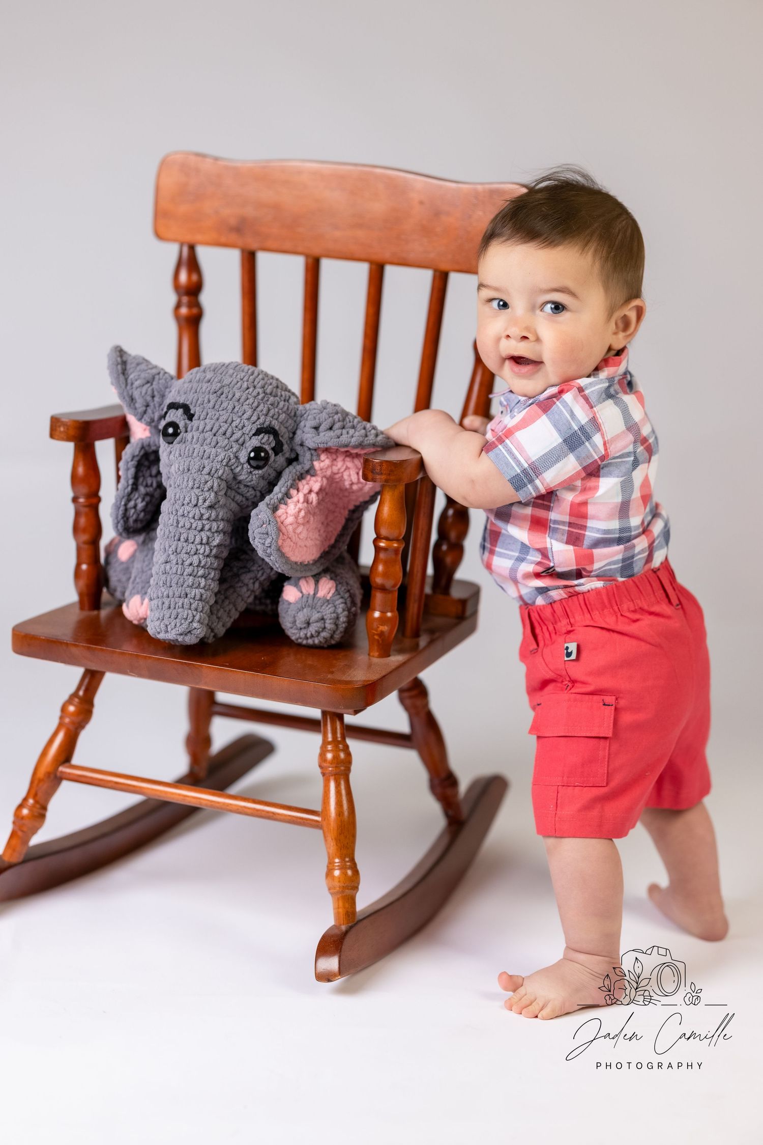A toddler in red shorts stands beside a rocking chair with a plush elephant toy.