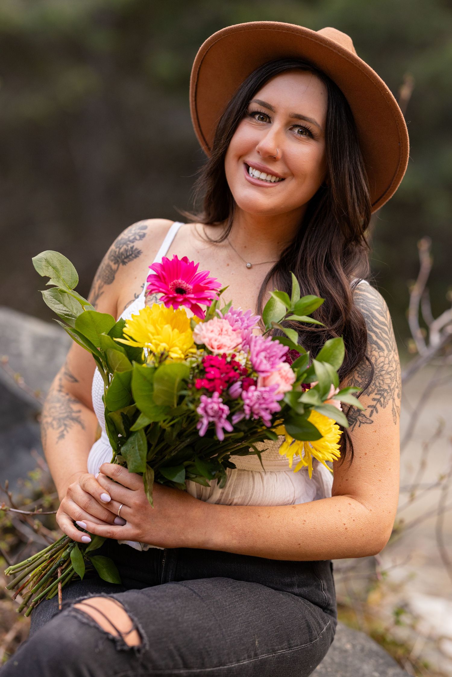 A person in a brown hat holds a colorful bouquet of gerbera daisies and chrysanthemums while sitting outdoors.