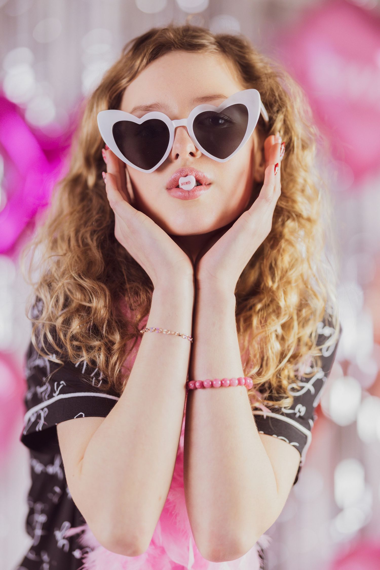 A person with curly blonde hair wearing heart-shaped sunglasses poses playfully against a pink bokeh background.