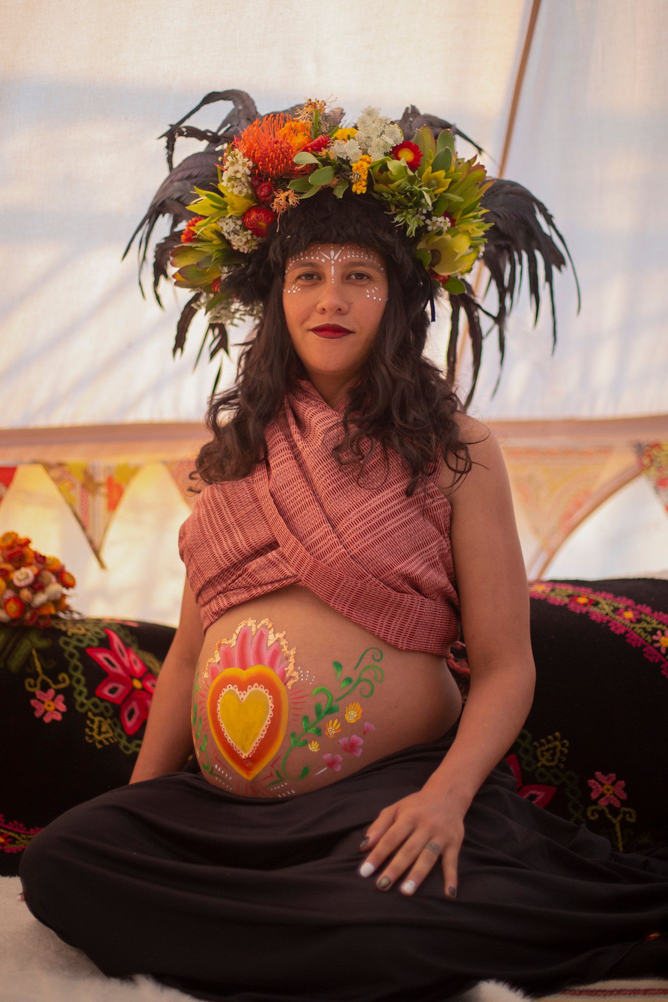 Bohemian maternity portrait with feathered floral crown and painted belly against bunting.