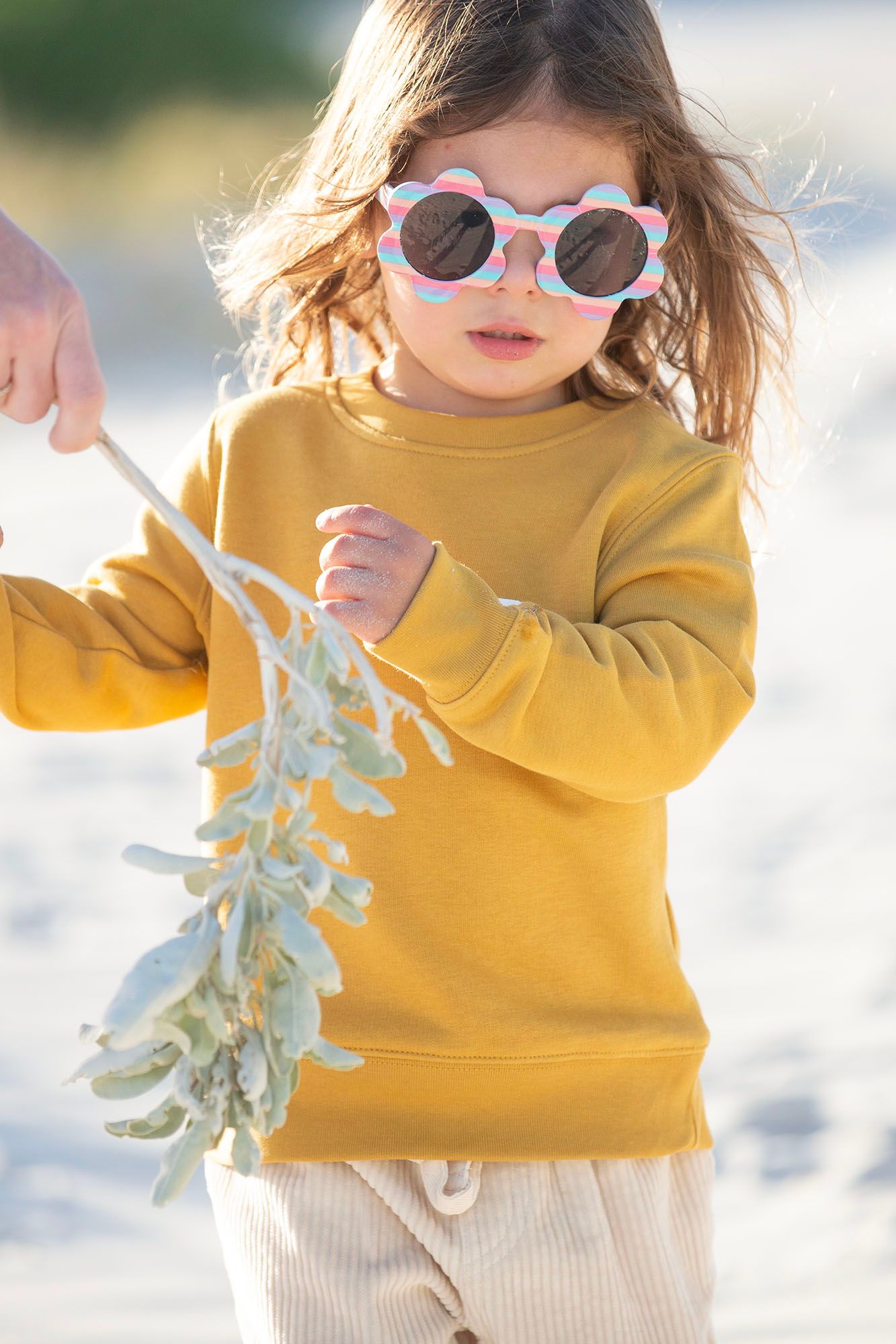 Child wearing sunglasses and yellow sweater playing with leaves in sunlight.
