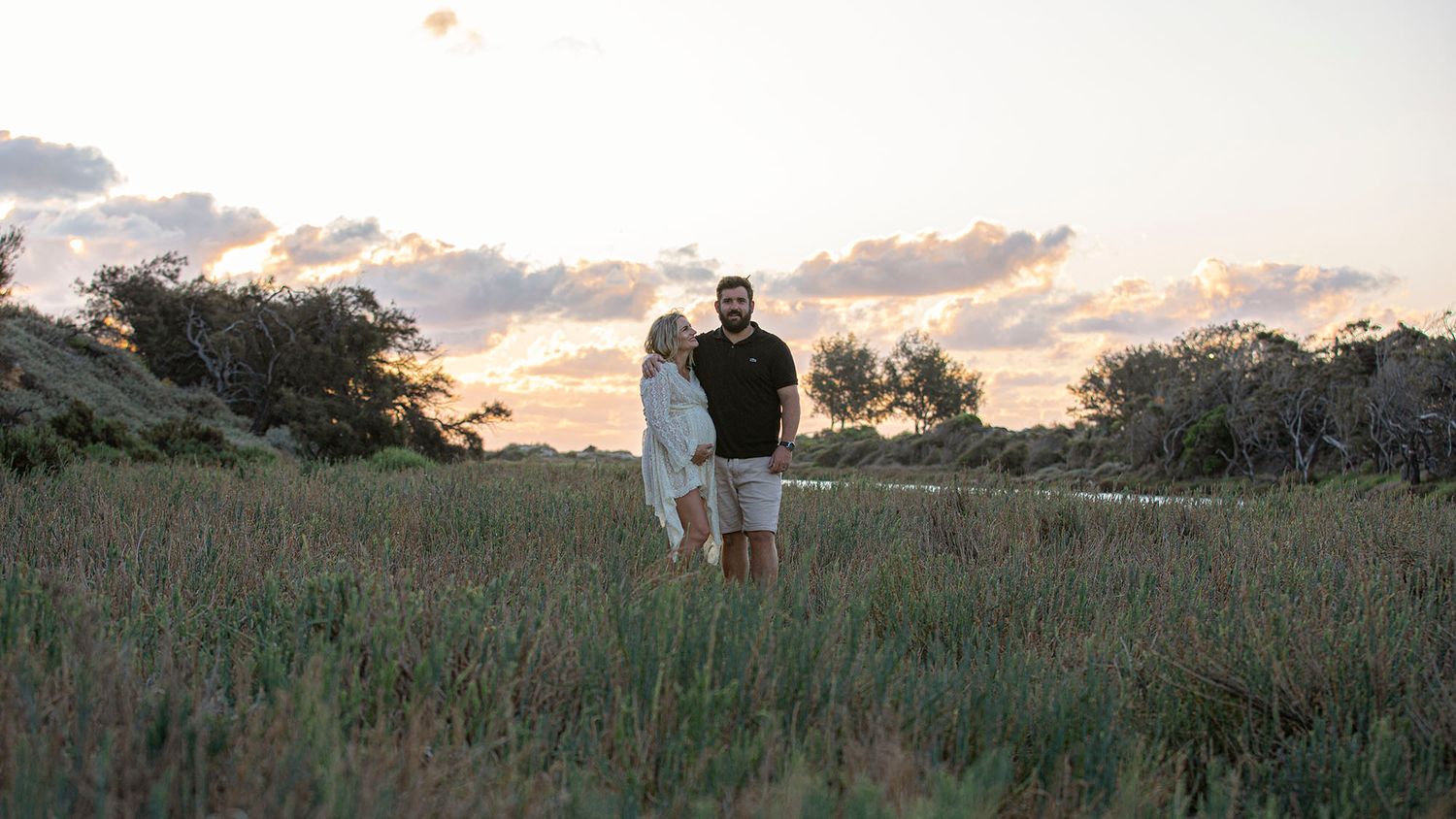 Two people stand together in a grassy field at sunset with trees silhouetted in the background.