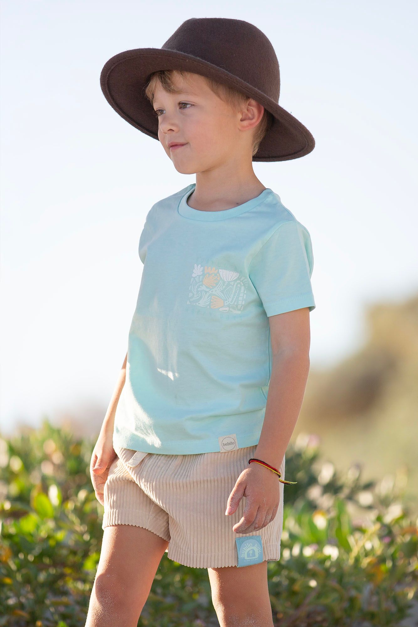 Child wearing light blue shirt, beige shorts and brown sun hat outdoors.