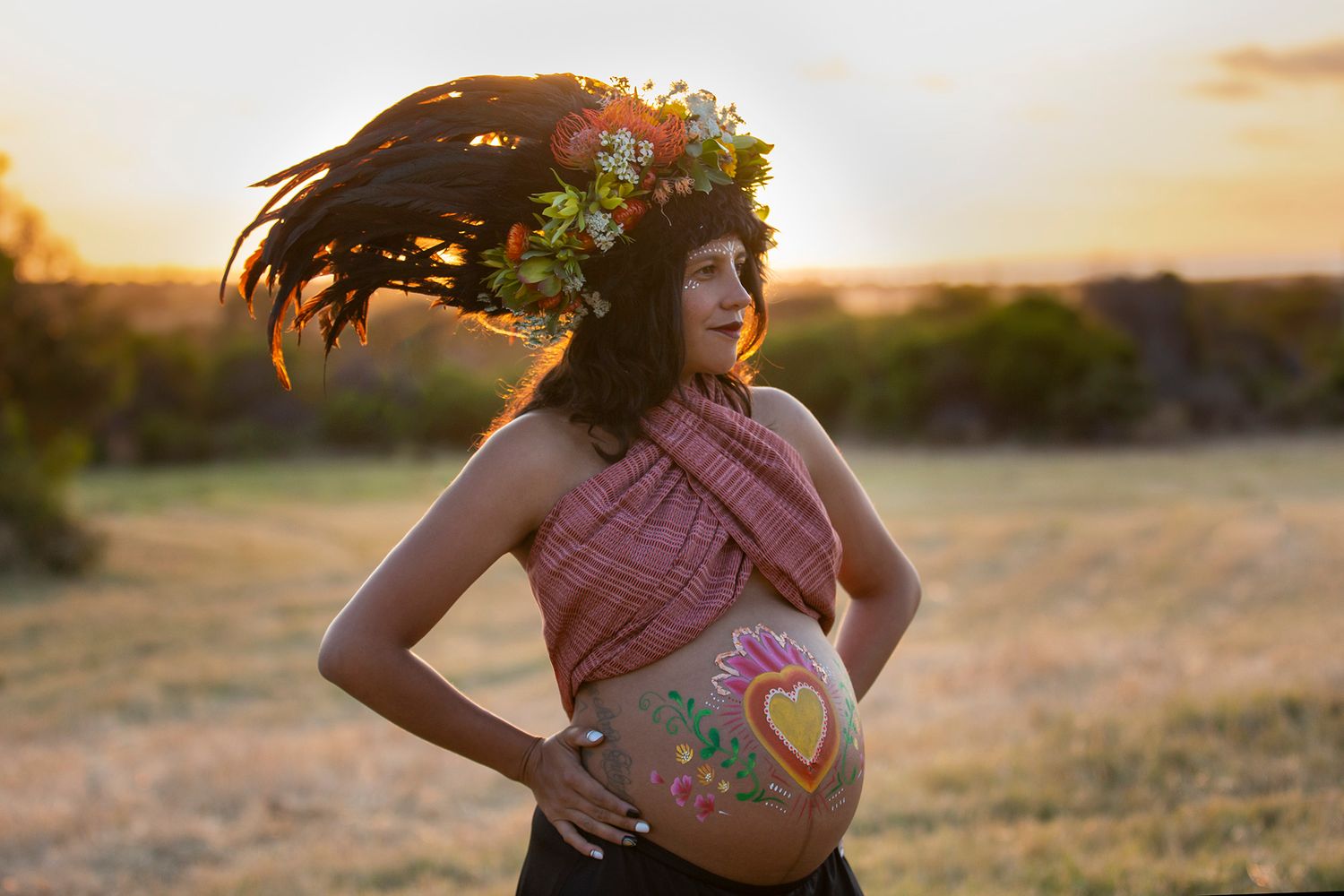 Expectant mother in pink top with floral belly art and feathered crown at sunset.