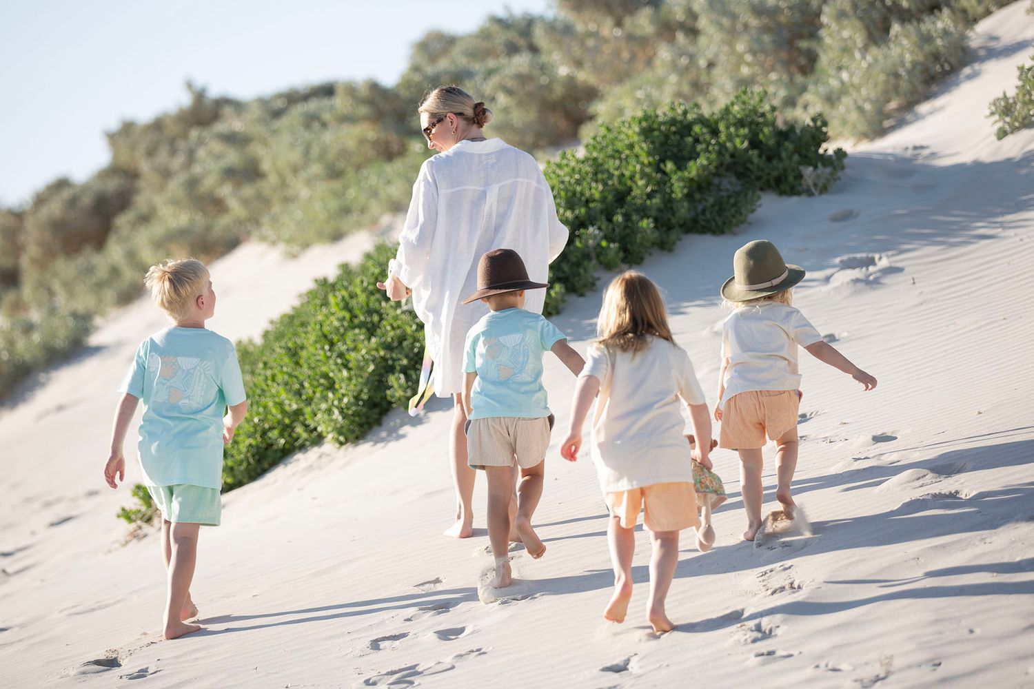 Small kids walking along a sandy beach path with plants and ocean in the background.