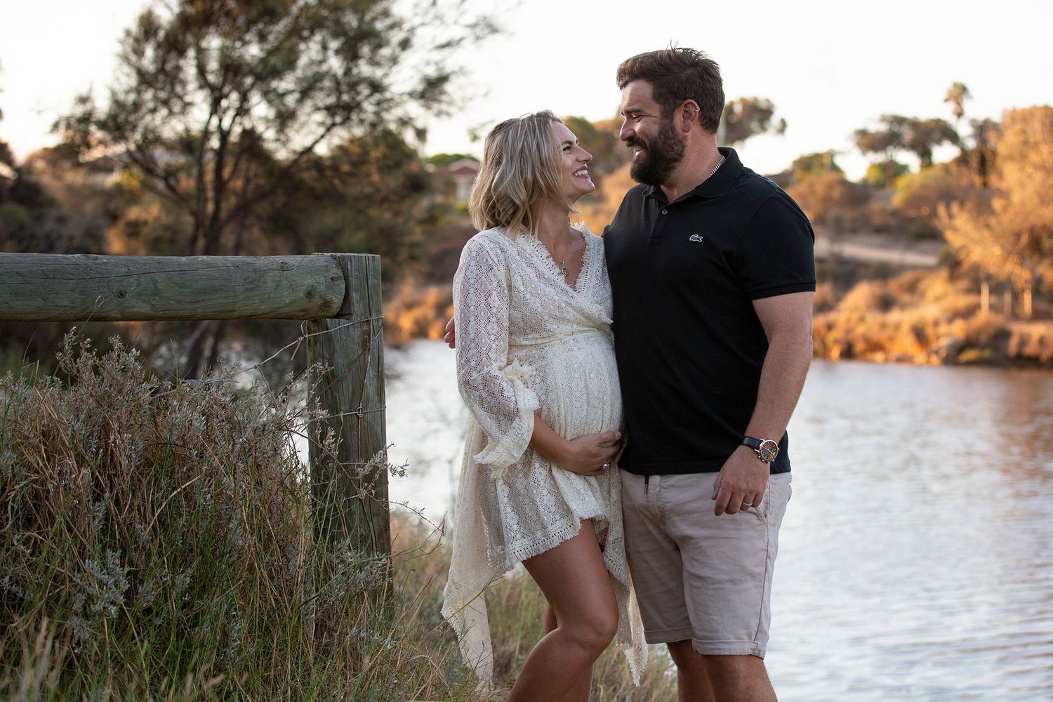 Romantic couple embraces by the water's edge during a golden sunset while wearing white and neutral toned outfits.