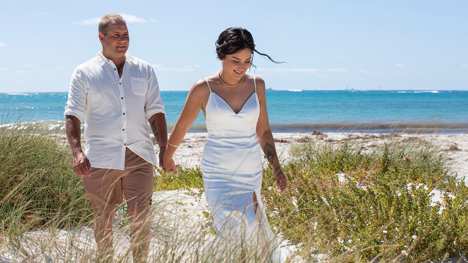 Couple in white clothing strolls along scenic beachfront path with turquoise ocean waters and sand dunes in view.
