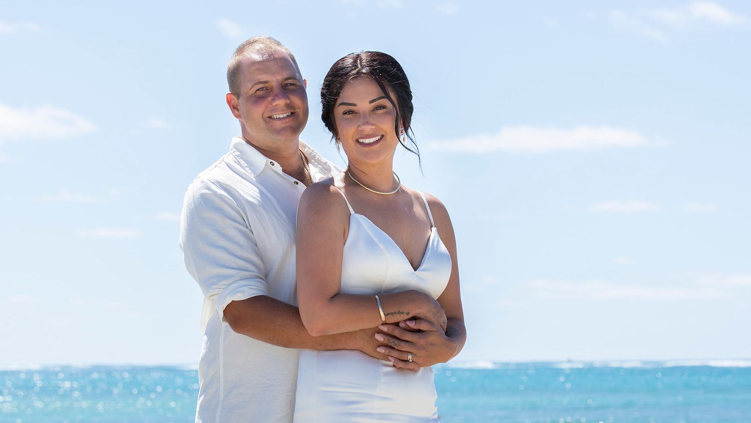 Happy couple embraces on tropical beach with bright blue ocean and sky in background during sunny day.