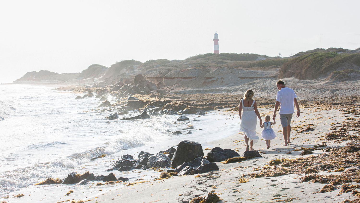 Family walking along rocky coastline with lighthouse in background.