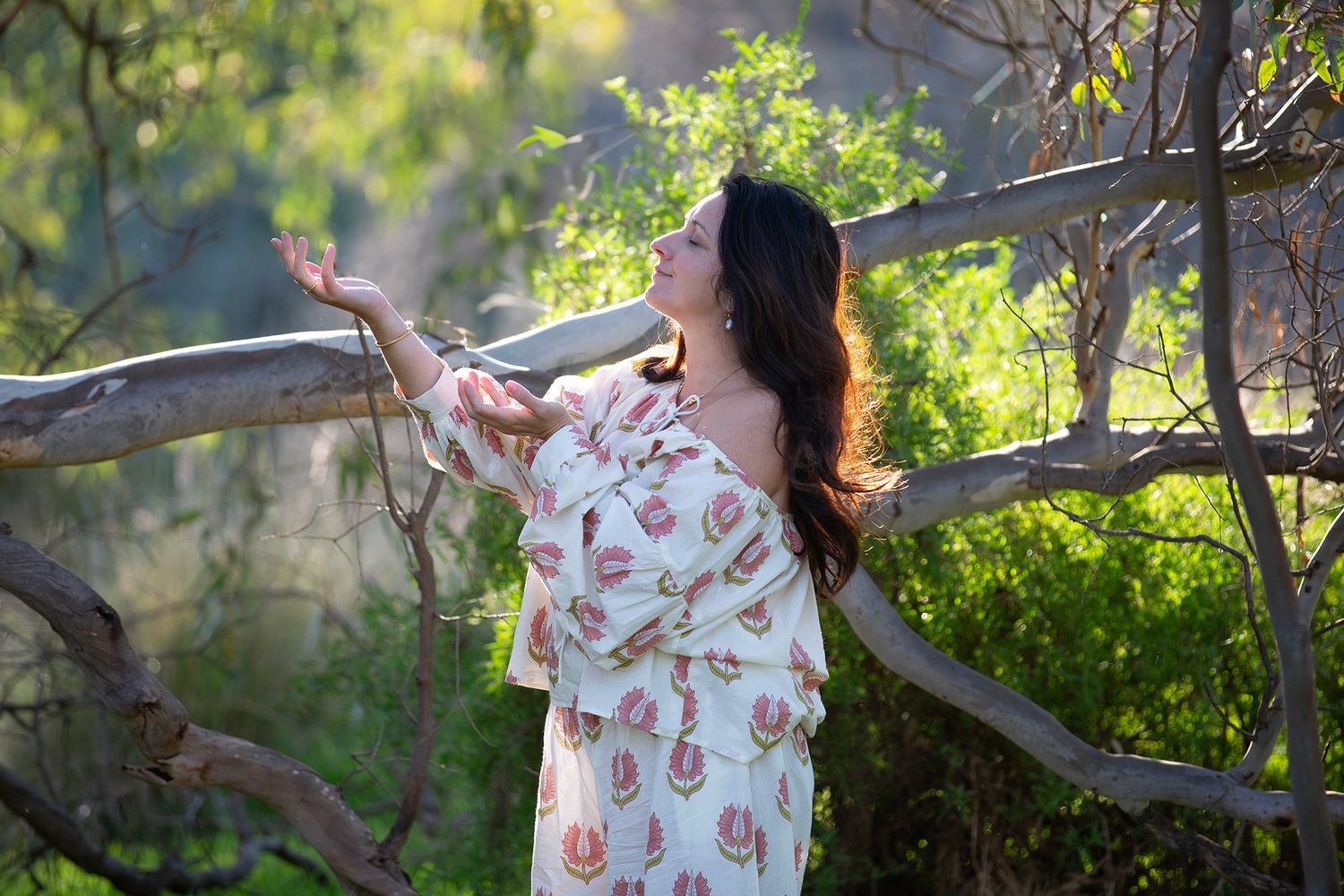 Person in floral dress standing among tree branches in sunlit garden setting.