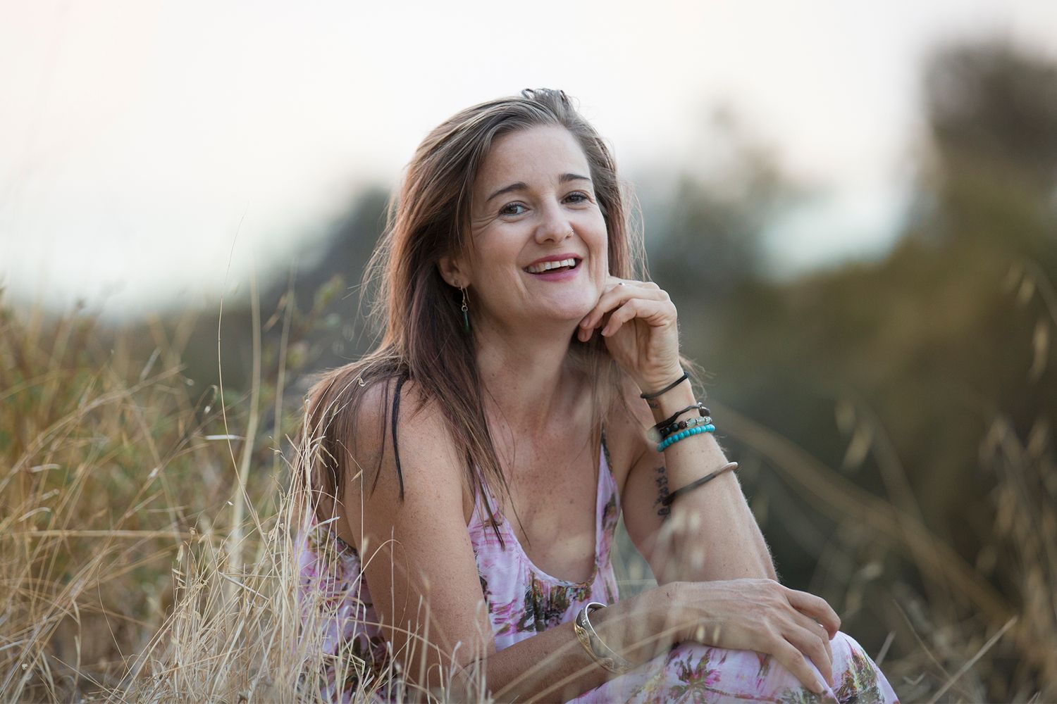 Woman laughing while sitting in a field of tall grass at sunset wearing a floral dress.