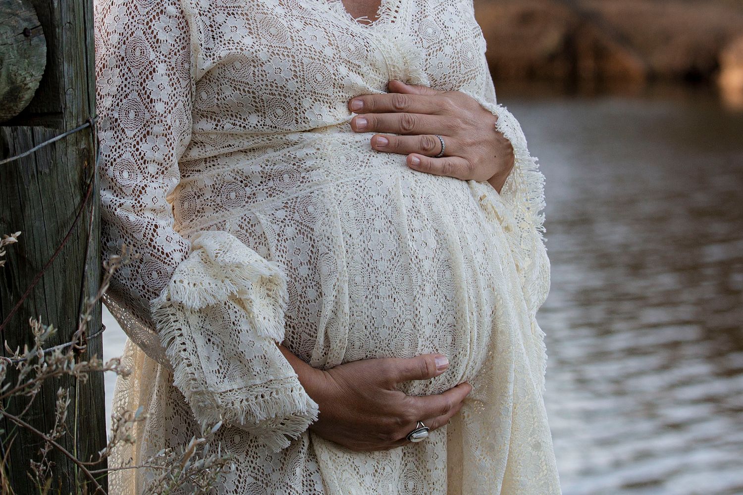 A pregnant woman wearing a delicate lace maternity dress stands in soft natural light during a maternity photoshoot.