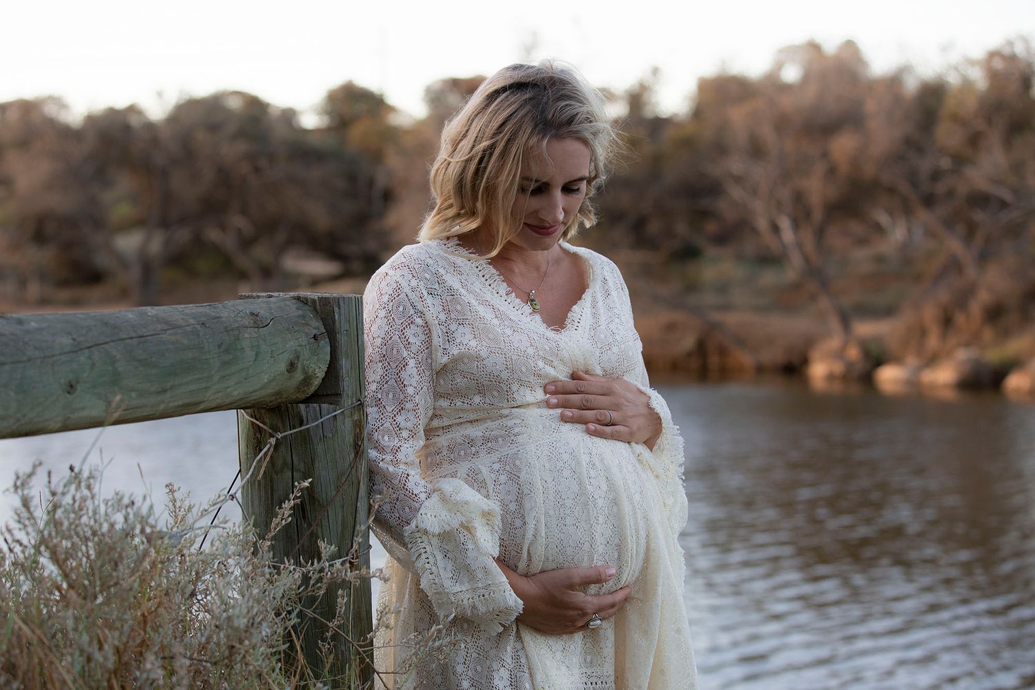 Ethereal maternity photo session by tranquil lake with woman in delicate white lace gown at dusk.