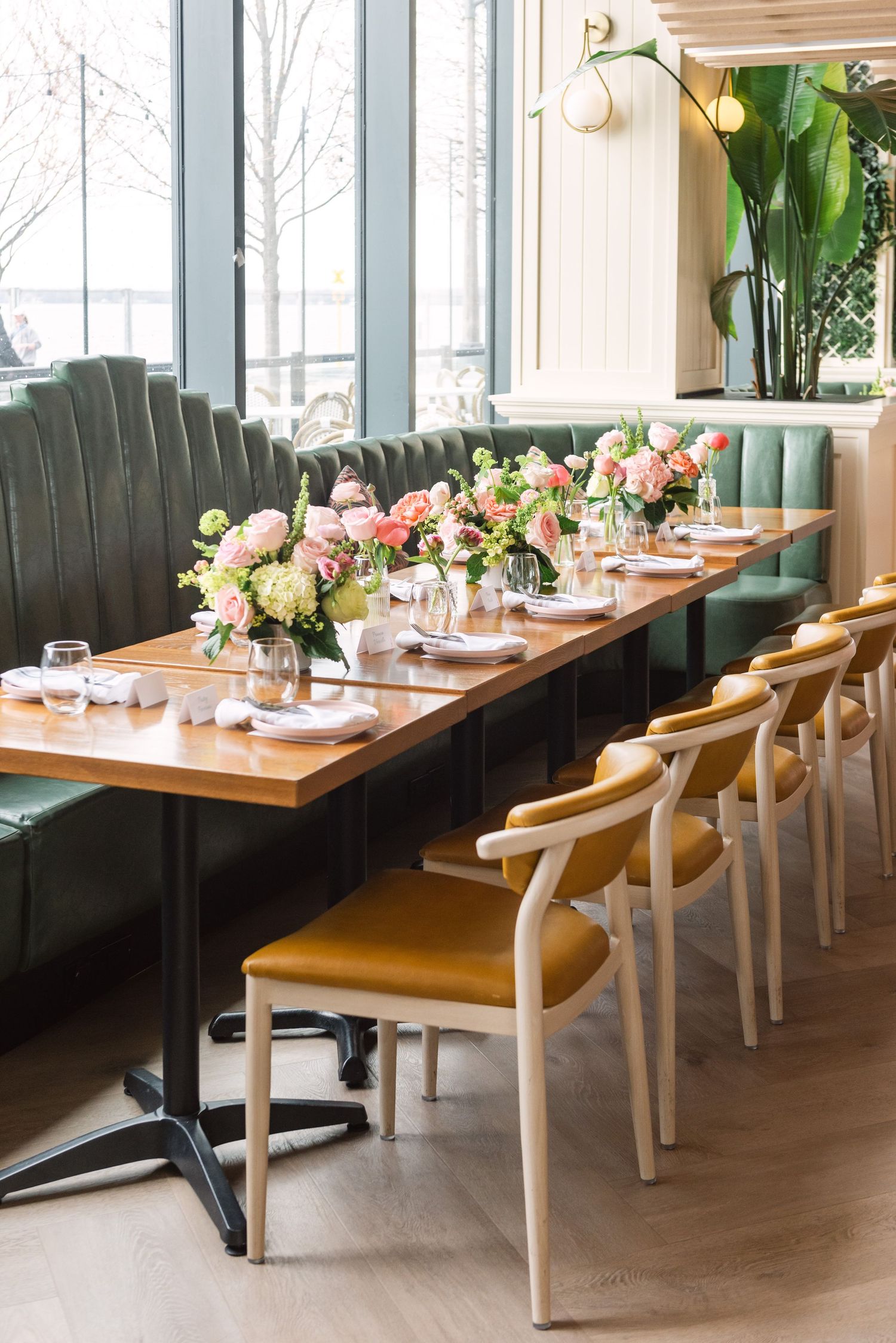 Elegant restaurant dining setup with green velvet banquette, wooden table, yellow chairs, and pink floral centerpieces.