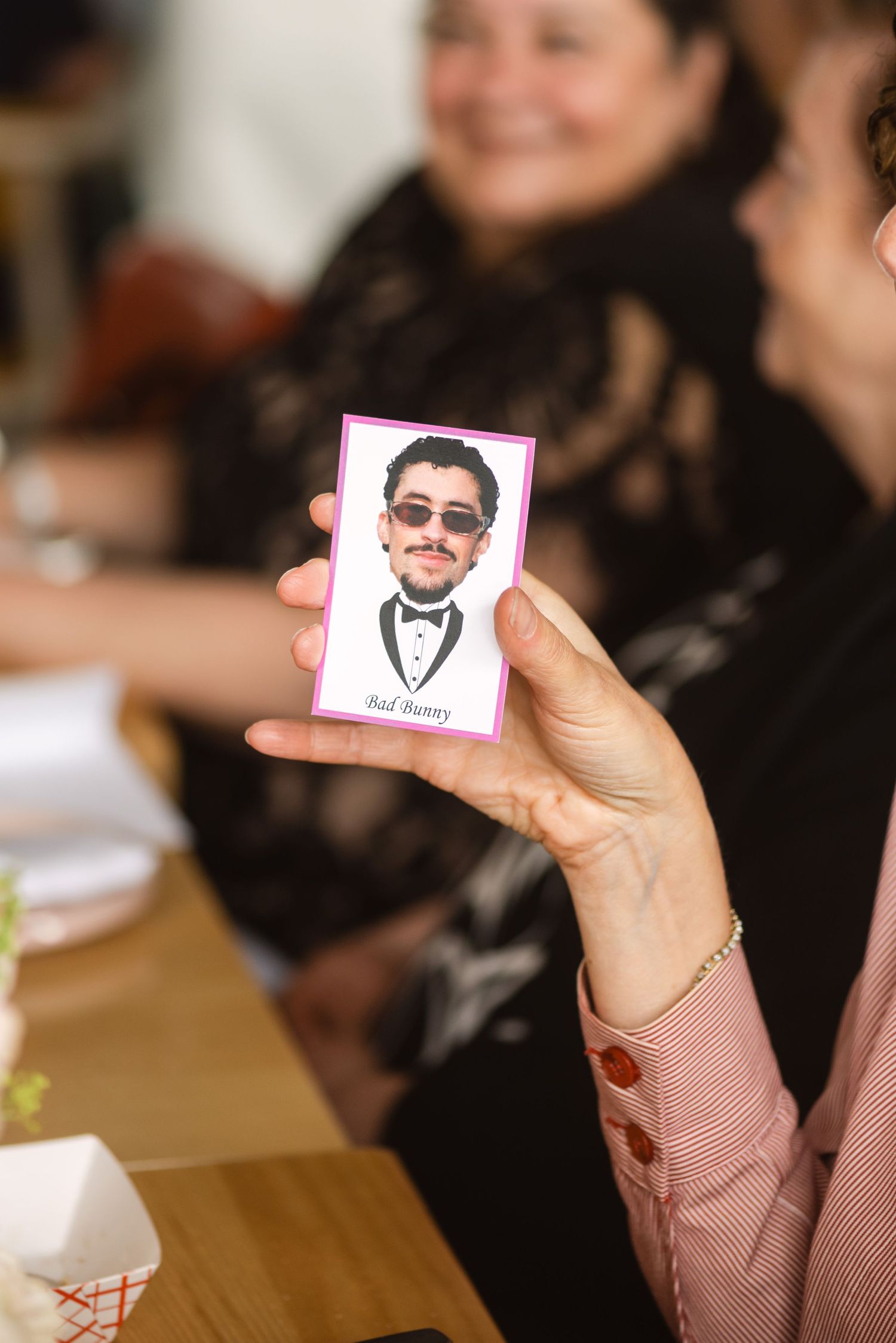 Hand holding a small photo card with a man in sunglasses and tuxedo, labeled Bad Bunny, at a social gathering.