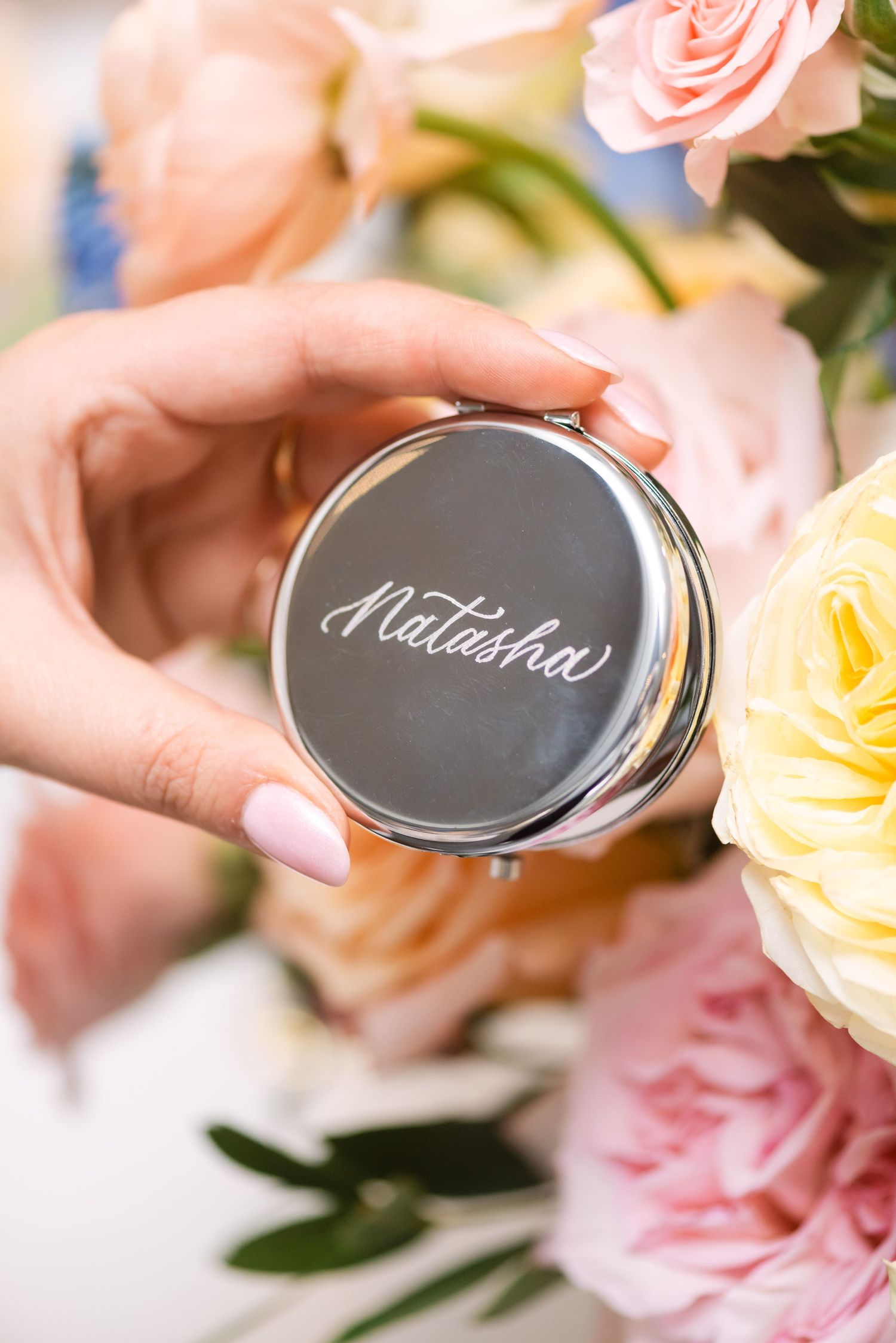 Hands holding a personalized silver compact mirror engraved with the name Natasha, surrounded by pastel wedding flowers.