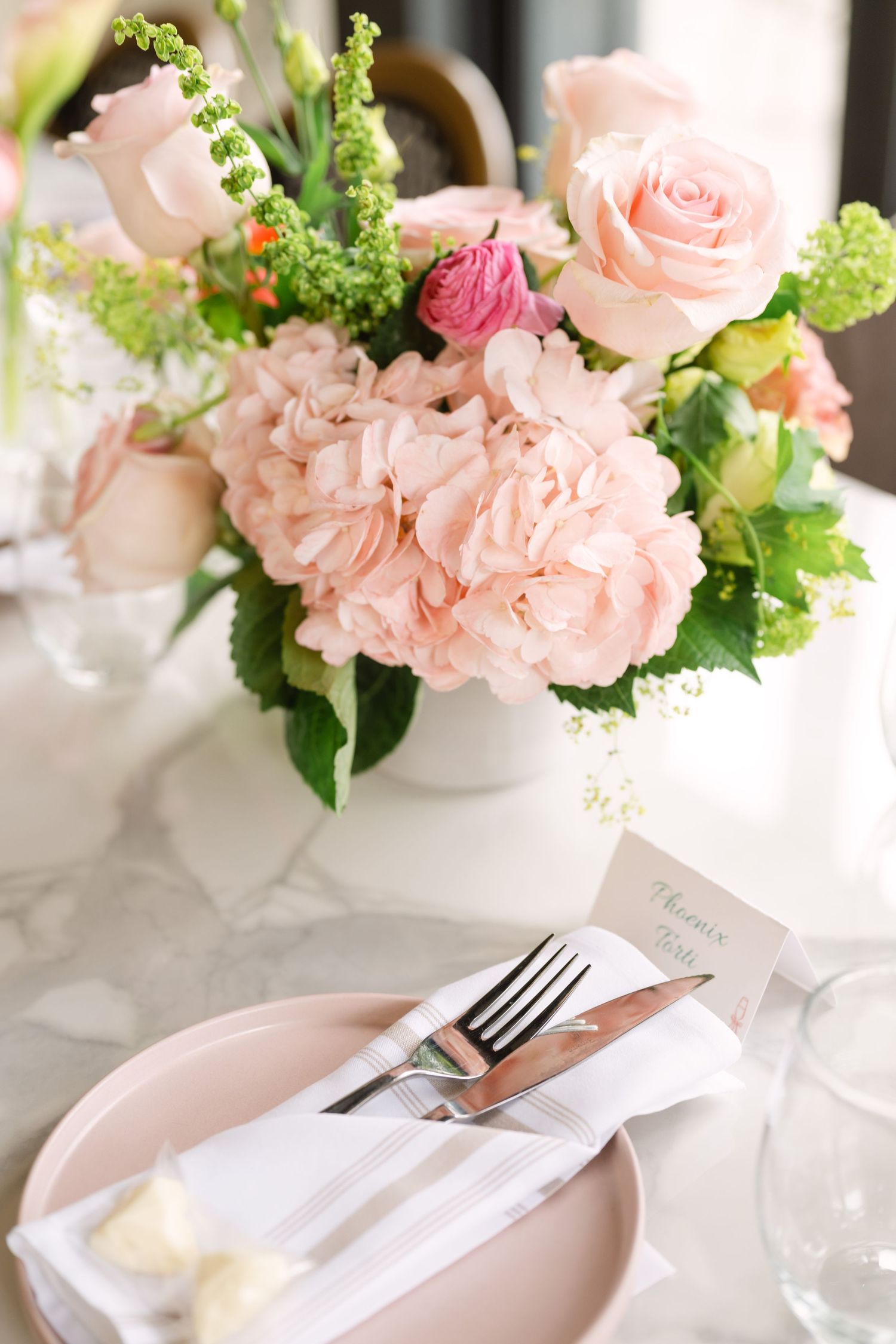 Elegant wedding table setting with blush pink hydrangeas and roses in white vase on marble table with pink plate and silverware.