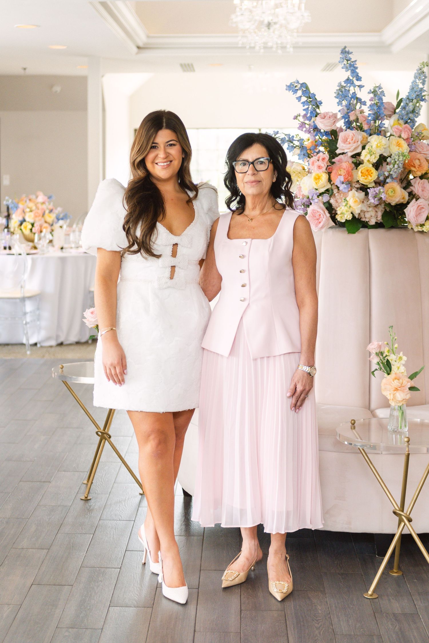 Two women pose together at an elegant bridal event, one in a white mini dress and one in a blush pink suit.