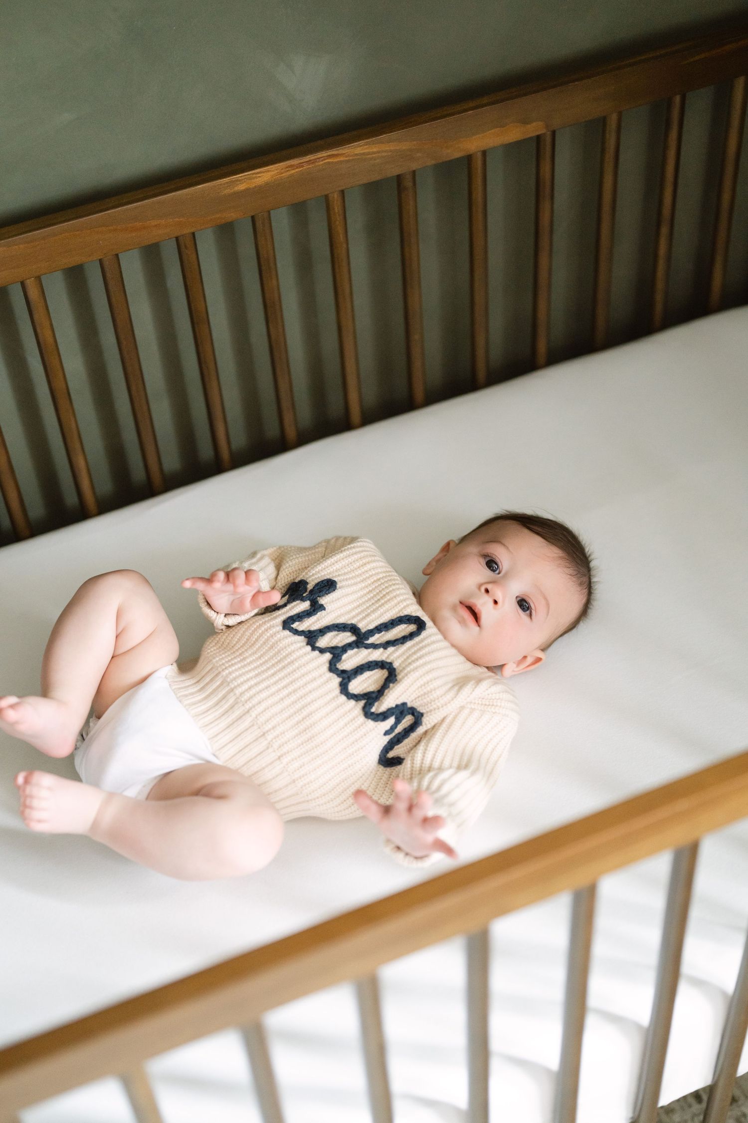 Baby wearing beige knitted onesie lying in wooden crib with white bedding.