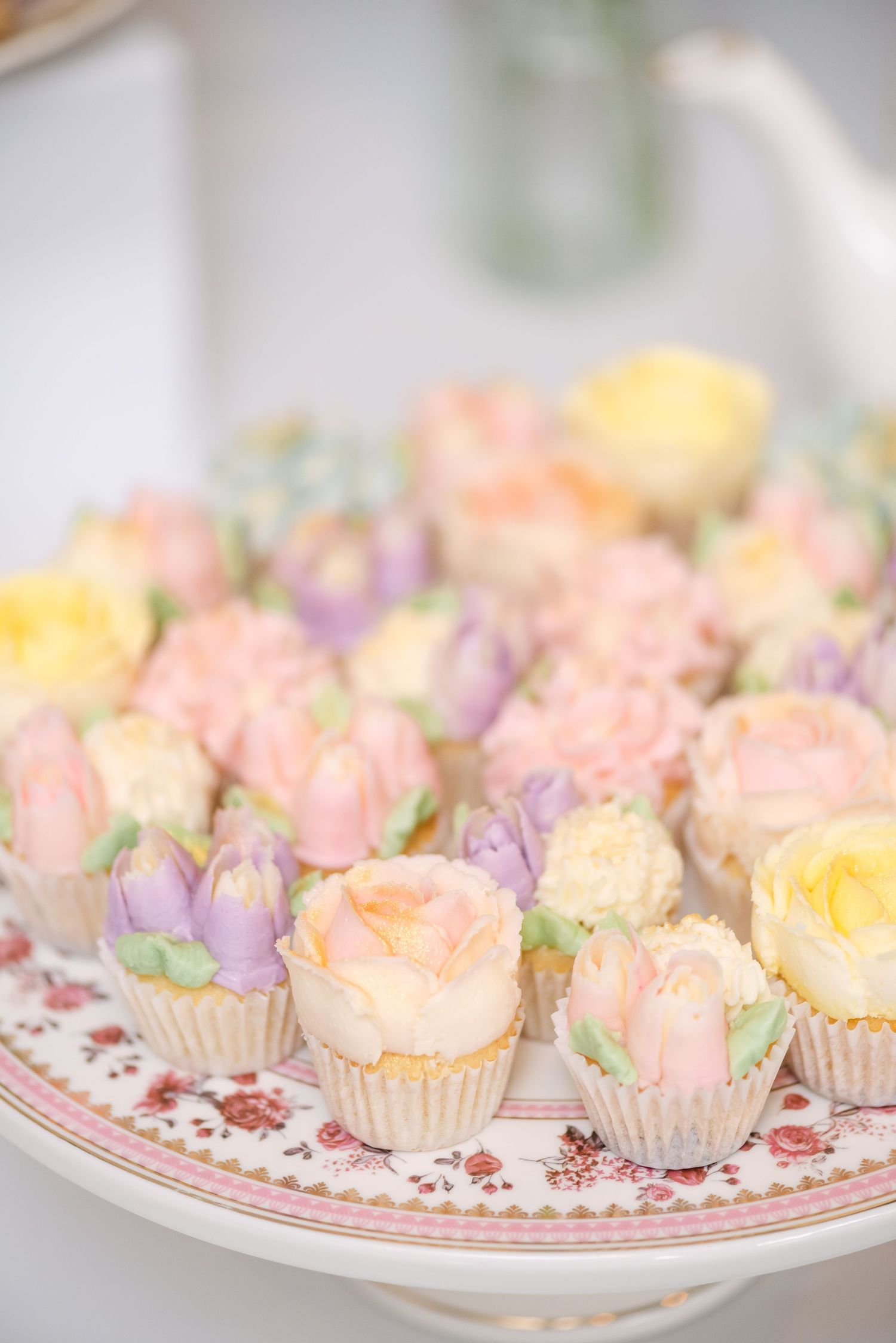 Elegant floral buttercream cupcakes in pink, lavender, and yellow displayed on a vintage rose-patterned cake stand.