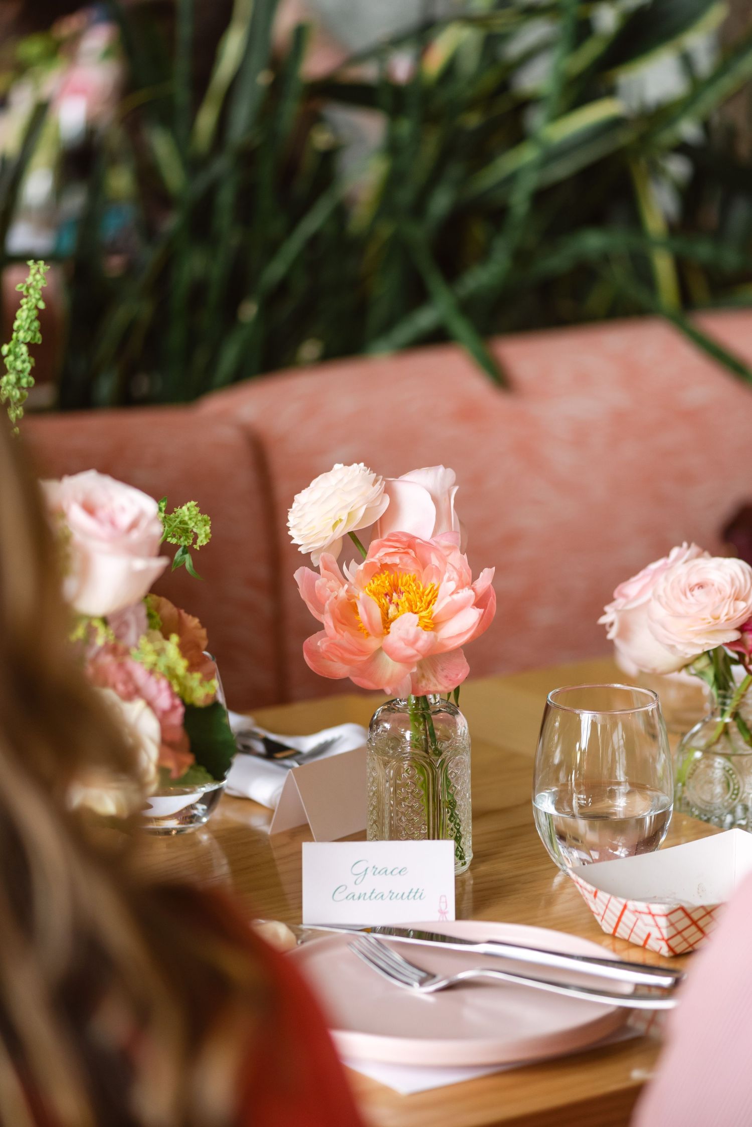Elegant wedding table setting with pink peonies and roses in glass vases, place card, and stemware on a wooden table.