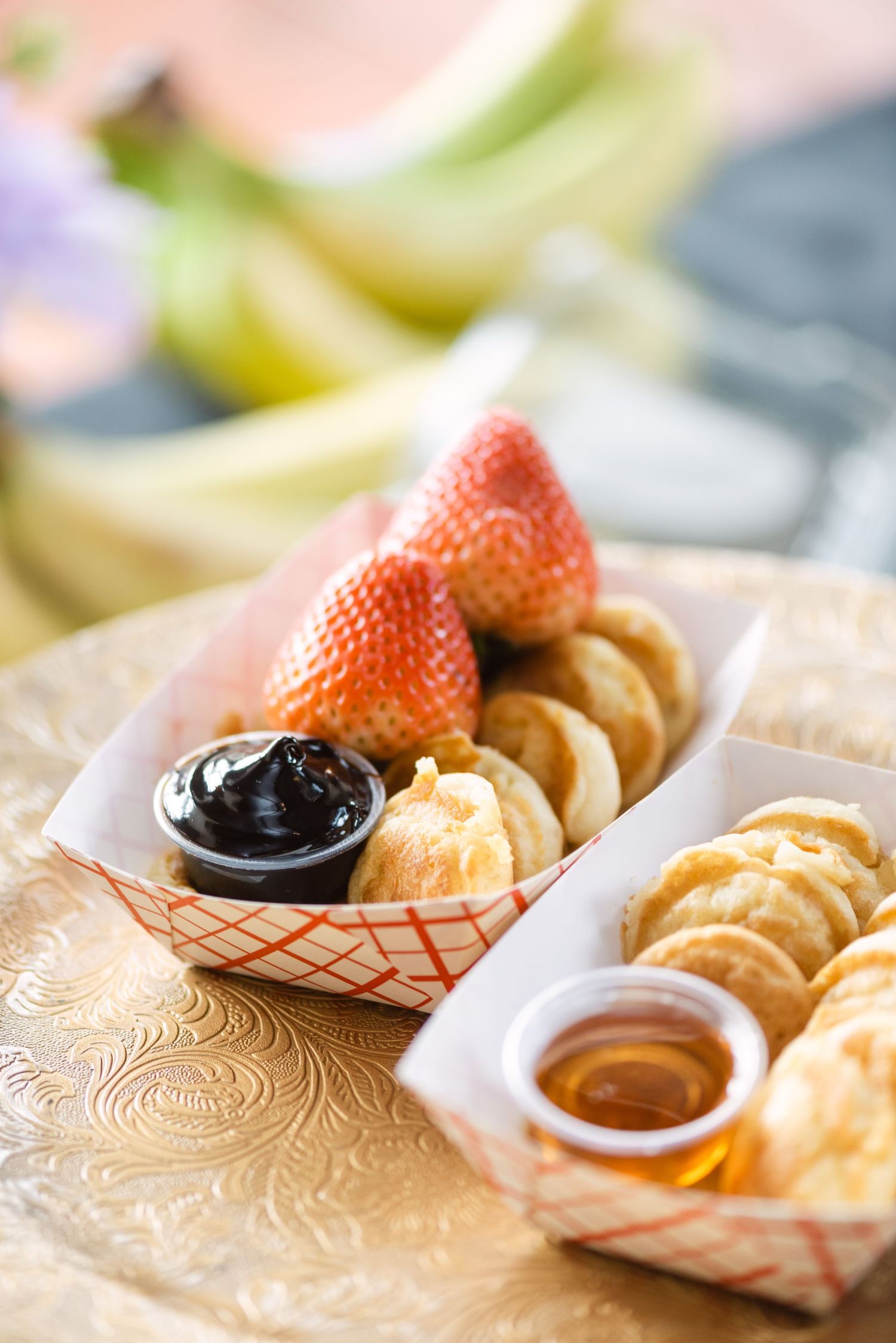 Mini pancakes and strawberries served in paper trays with chocolate and honey dipping sauces on a decorative gold plate.