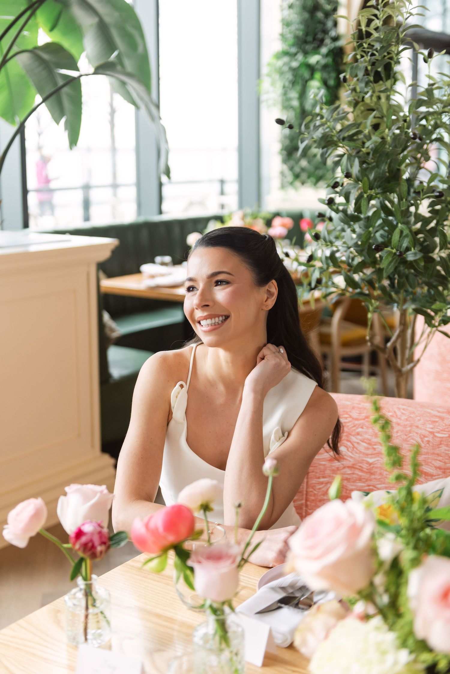 Smiling young woman in white dress sitting at a restaurant table with pink flowers and lush greenery.