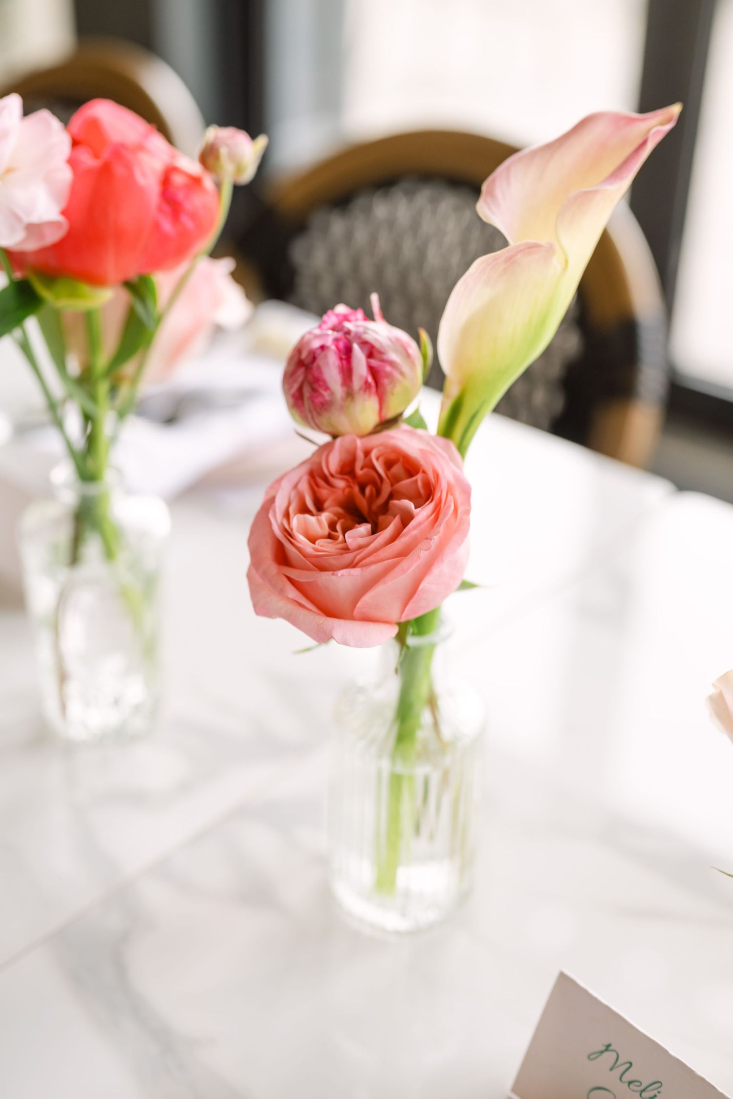 Delicate pink garden roses and calla lily stems in clear glass bud vases on white marble table at wedding reception.
