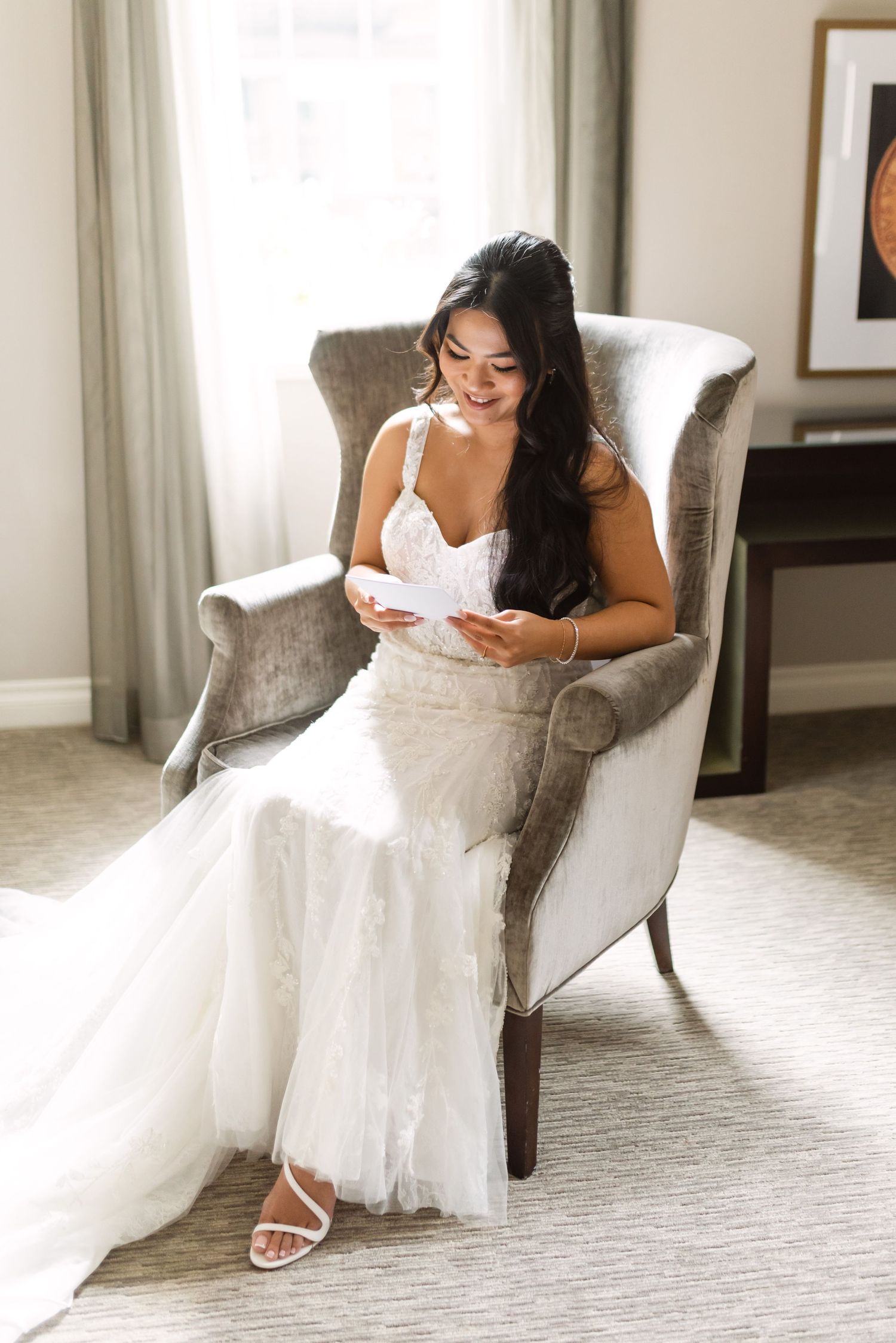 A bride in a white gown sits elegantly in a gray armchair while reading.