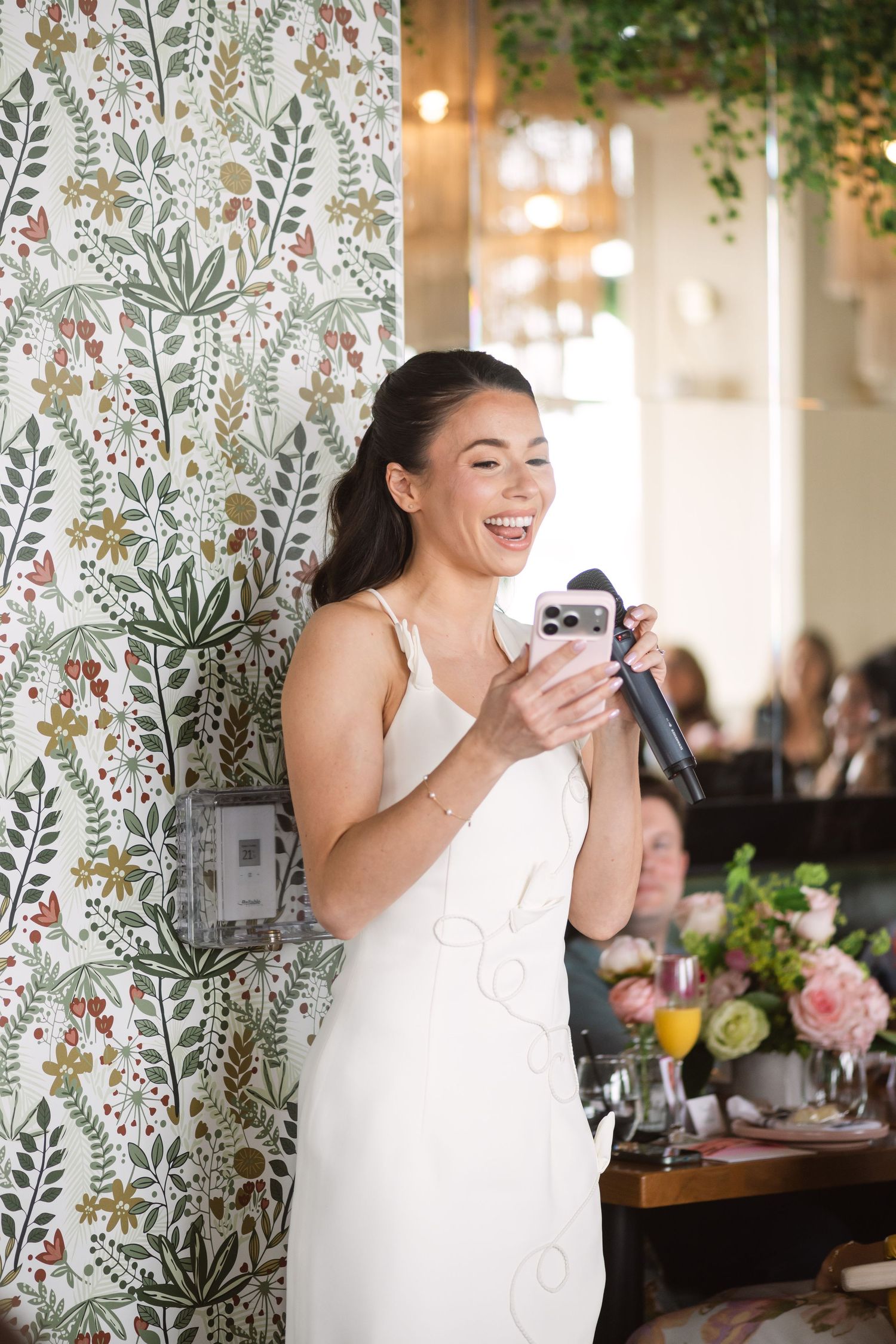 Bride in white dress laughing while giving speech with microphone at wedding reception.