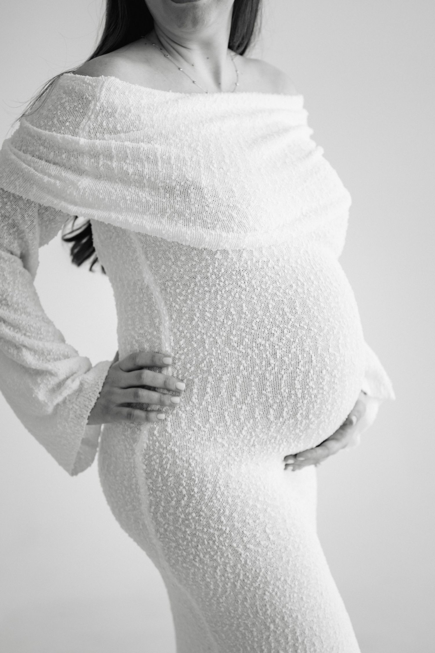 Black and white maternity photo showing a pregnant woman in a textured white dress with an off-shoulder wrap detail.