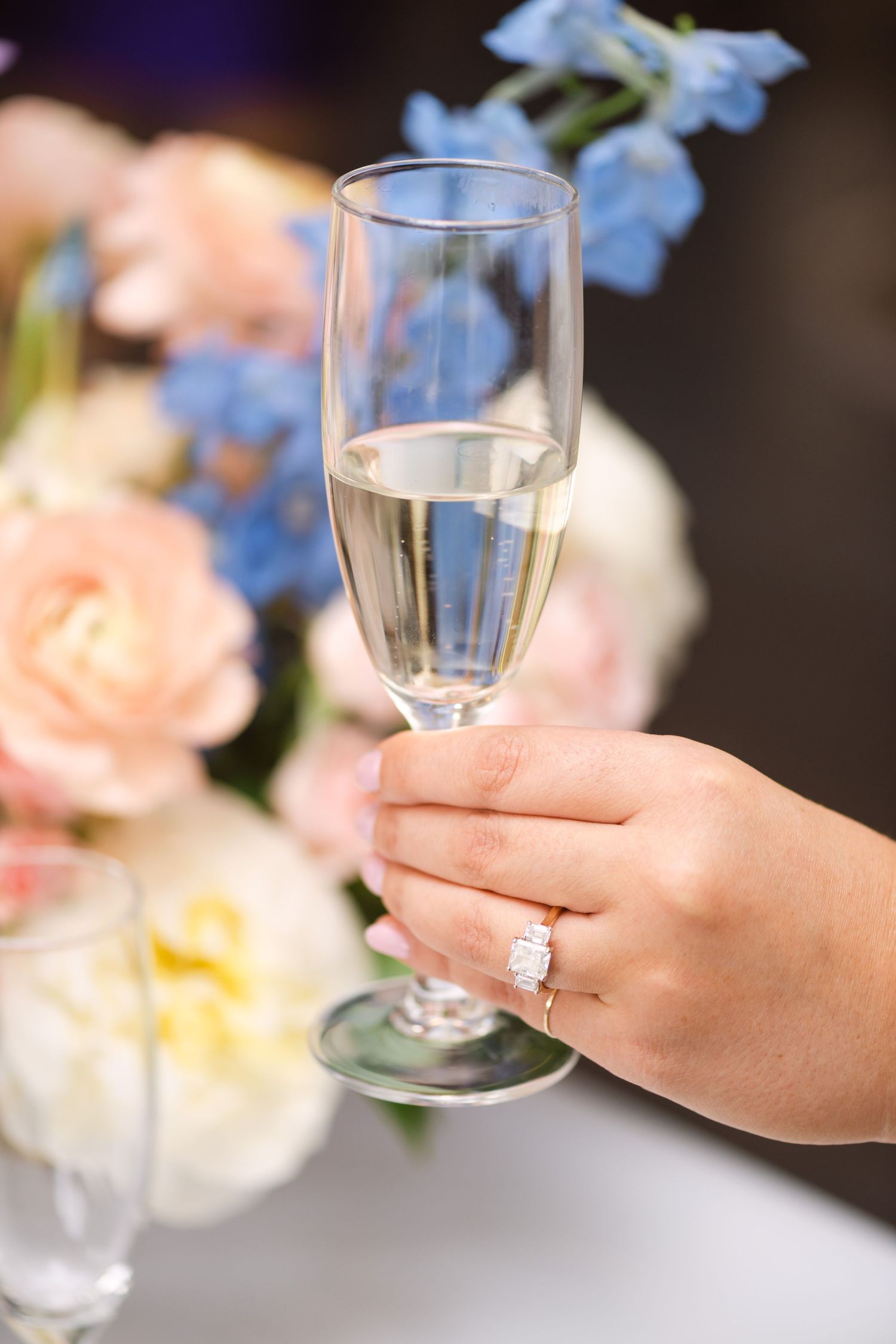 Hand holding champagne flute with diamond engagement ring, soft floral arrangement in pink and blue in background.