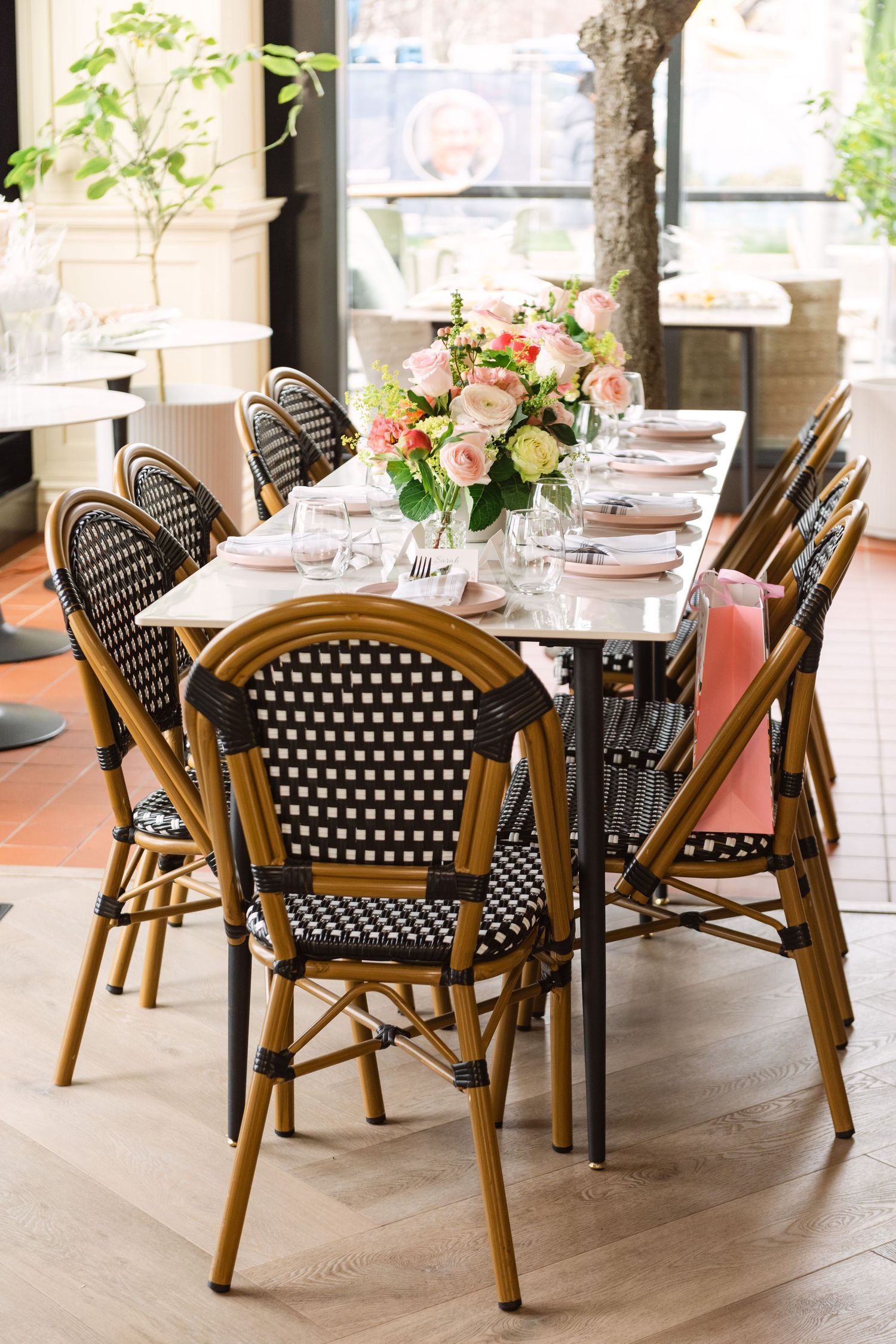 Elegant bistro table with black wicker chairs, pink floral centerpiece, and place settings in a bright restaurant.