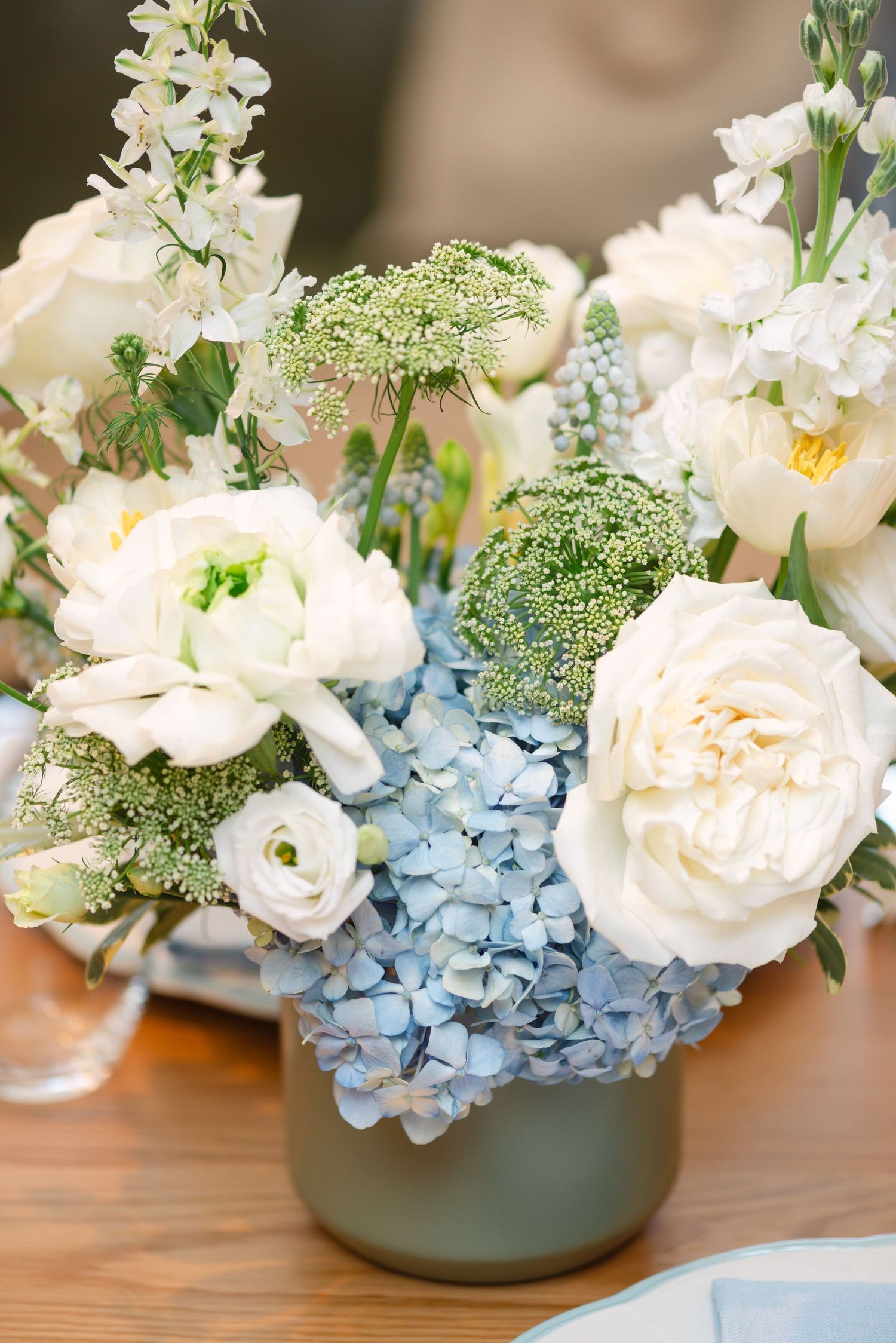 Elegant floral arrangement featuring soft blue hydrangeas and white roses in a gray ceramic vase on a wooden surface.
