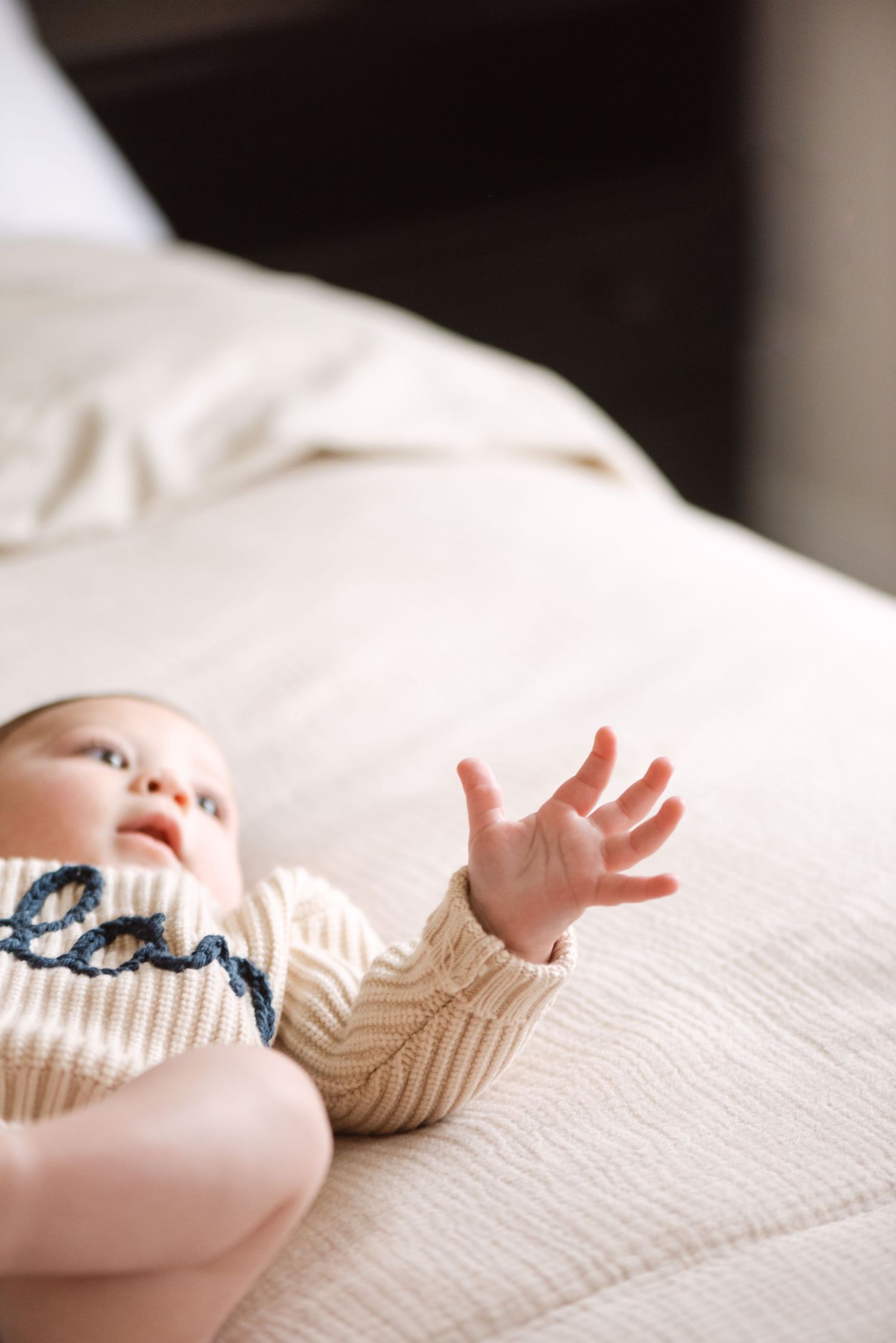 A baby's tiny hands reach upward while lying on a white textured blanket or surface.