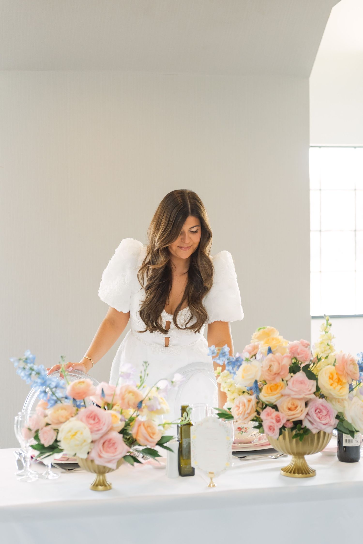 Woman in white dress arranging colorful floral centerpieces with pink, peach, and blue flowers on an elegant white table.