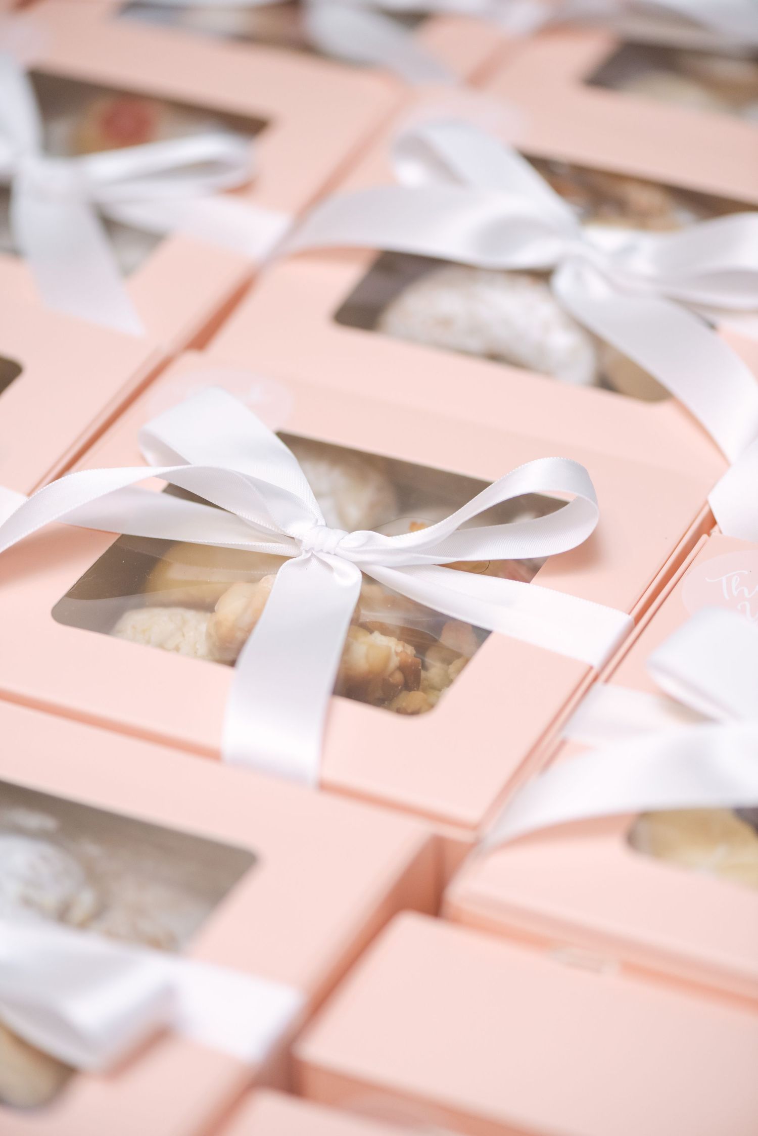 Pink gift boxes with clear windows and white satin bows, filled with assorted cookies, arranged in rows.