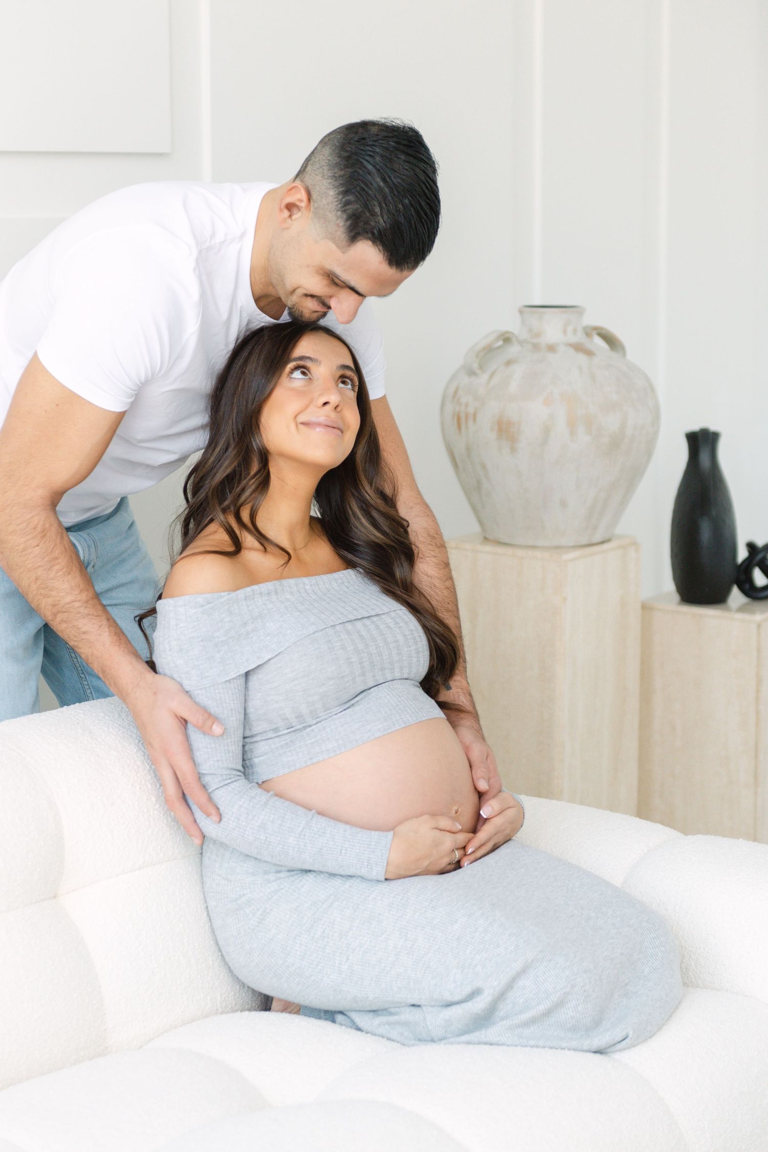Intimate maternity photos captured on white bedding with decorative vases in background.