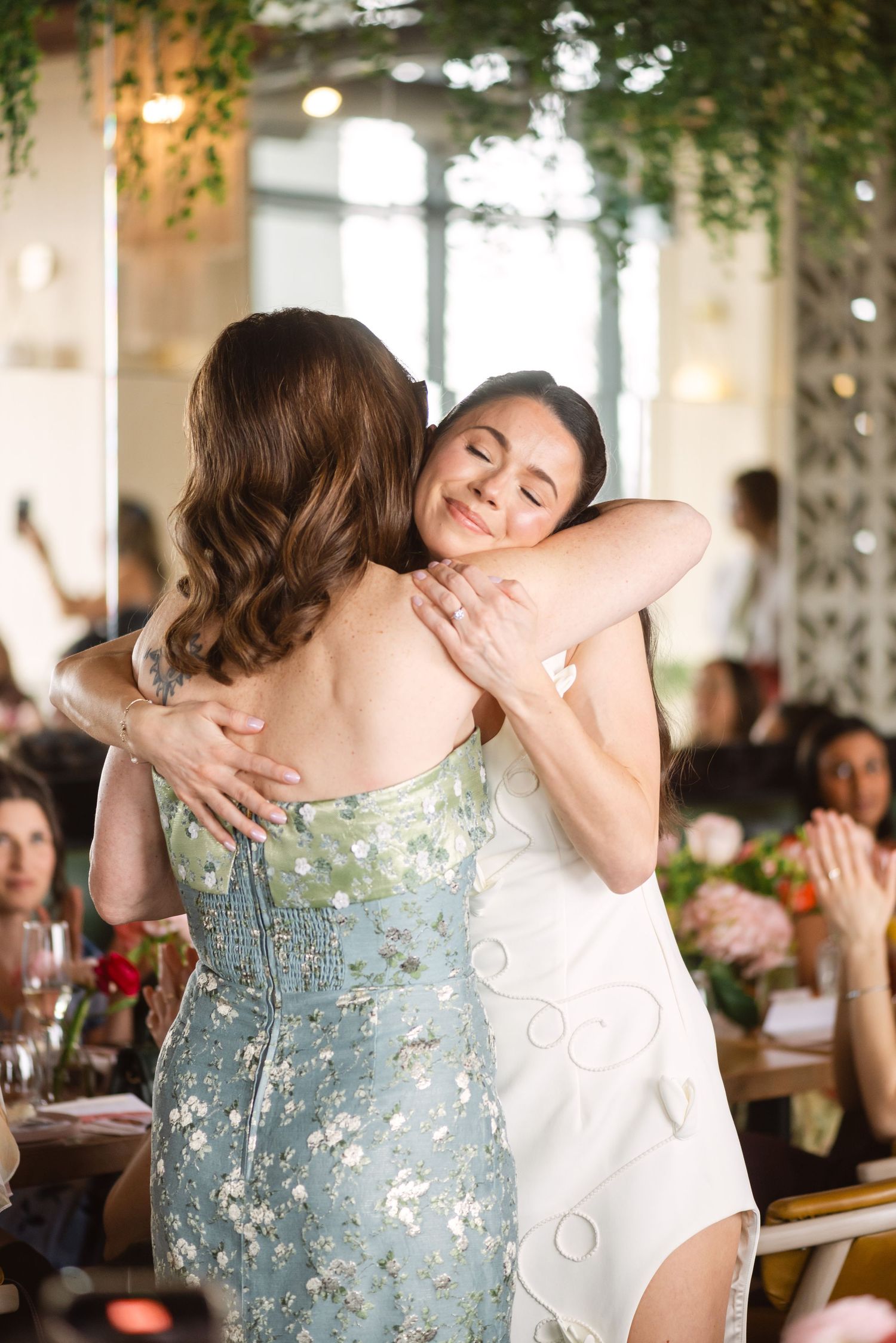 Two women share a warm hug at an elegant indoor event, one in a floral dress, smiling with eyes closed.