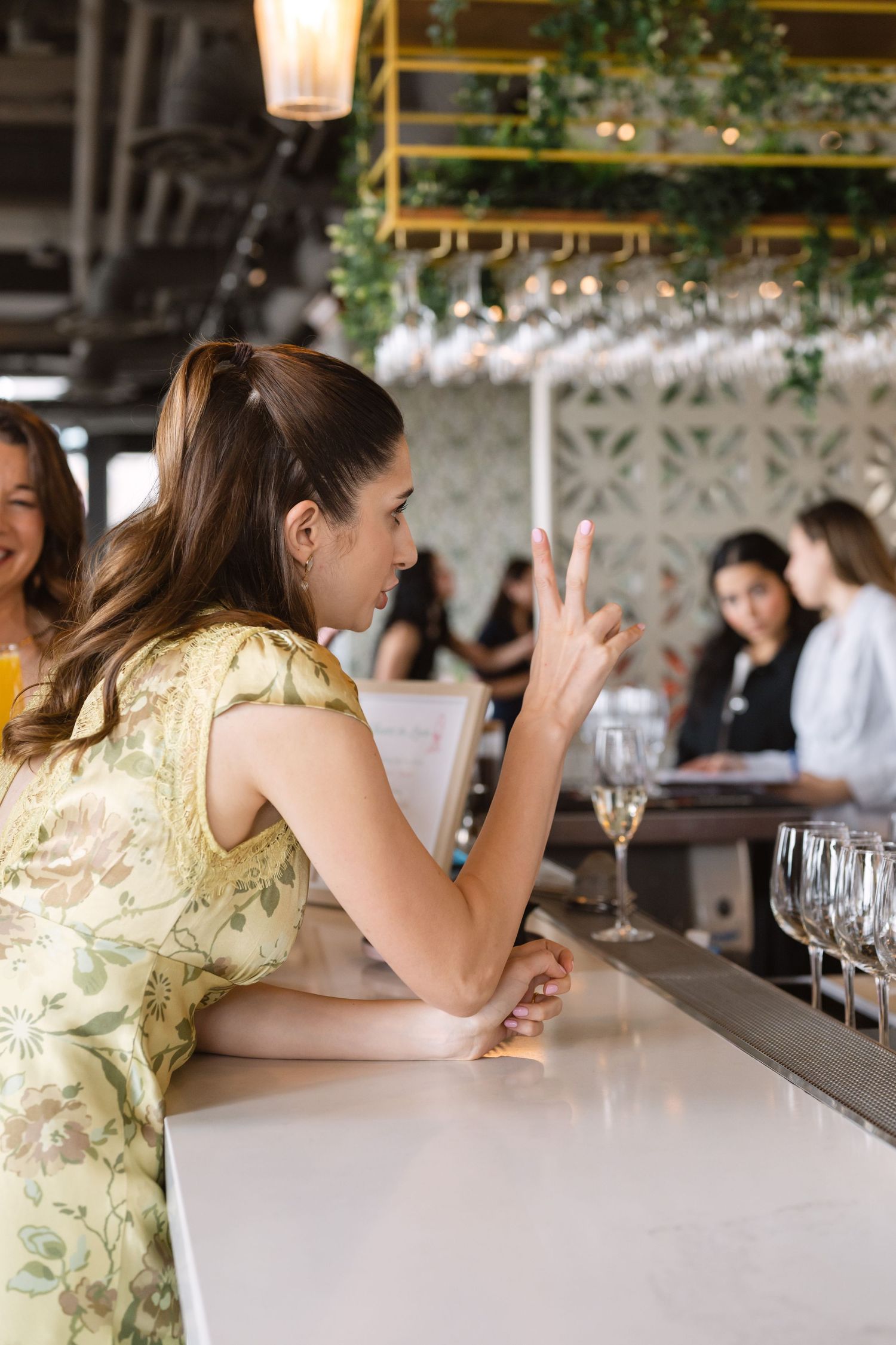 Young woman in yellow floral dress sitting at marble bar gesturing to bartender at upscale restaurant event.