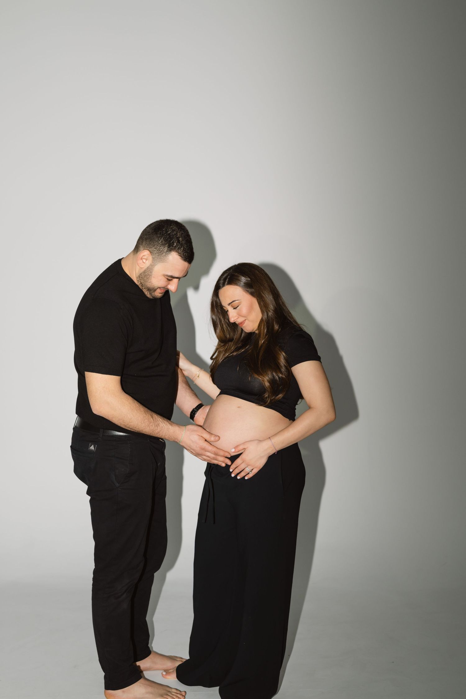 Couple in black clothing shares intimate moment during maternity photo shoot against white background.