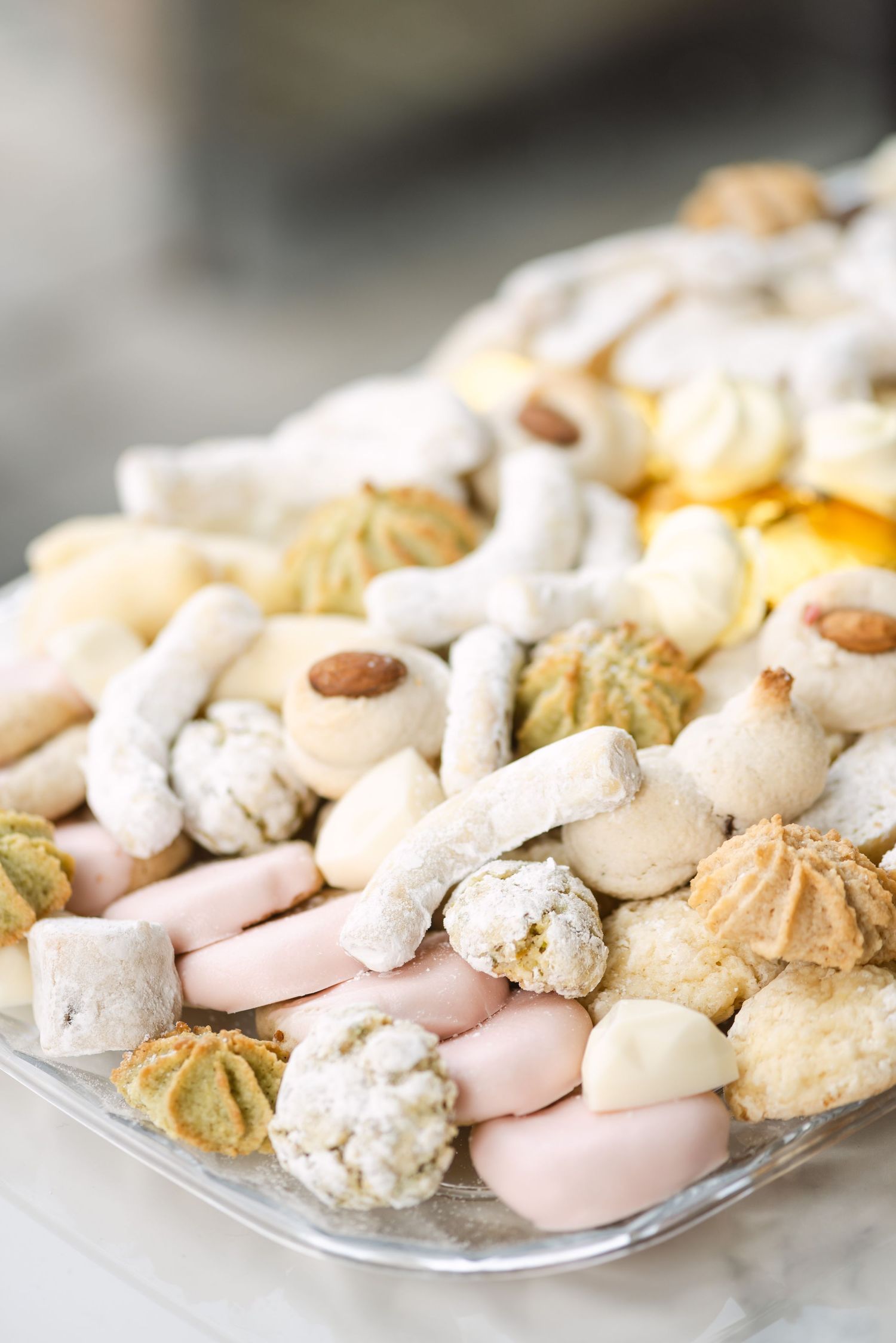 Assorted cookies and sweets on a glass plate, including powdered sugar treats, almond cookies, and pastel-colored confections.