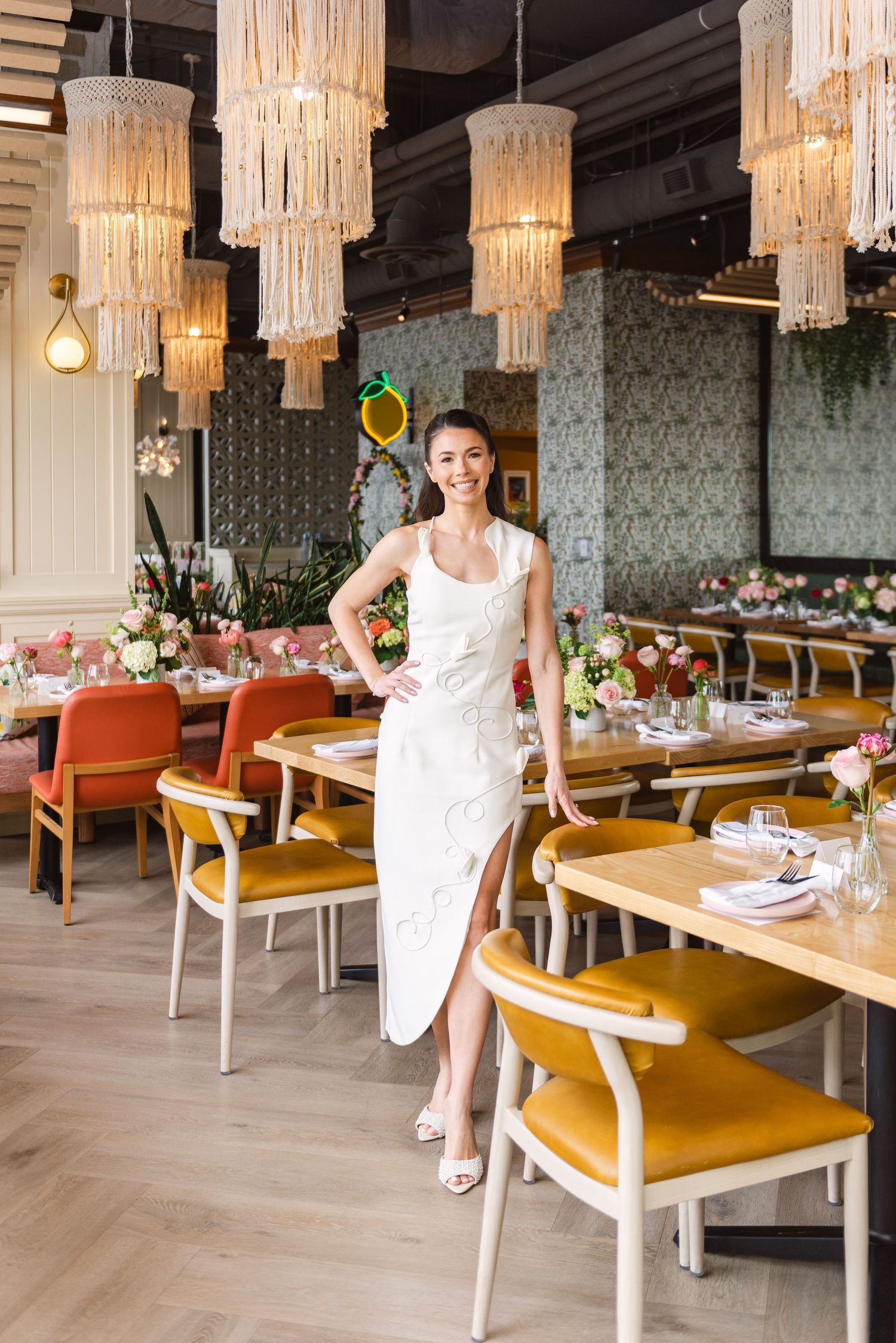 A woman in a white midi dress poses in an elegant restaurant with macrame chandeliers and colorful chairs.