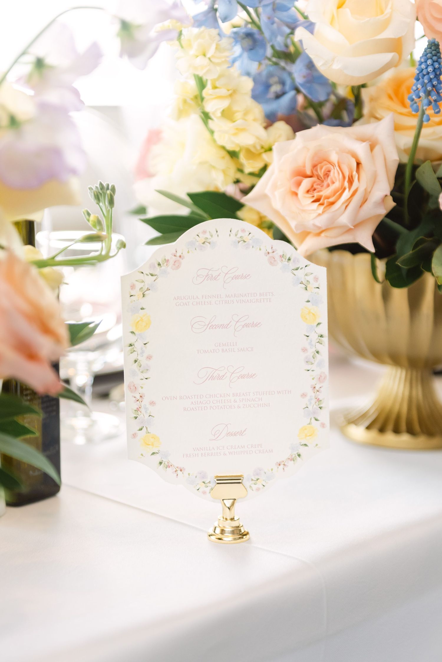 Elegant wedding menu card with floral border on gold stand, surrounded by peach roses and pastel flowers.
