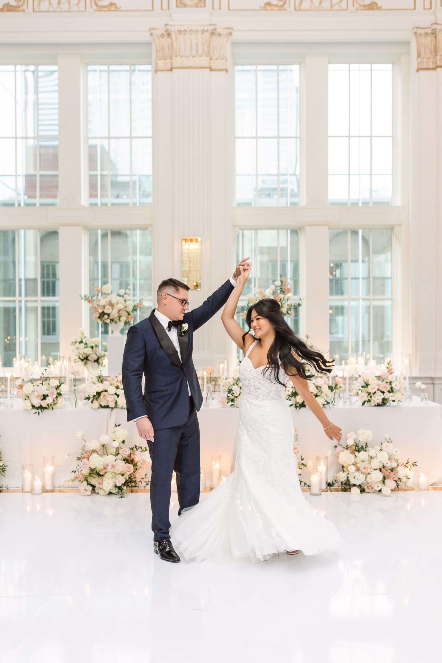 A couple dances in a beautifully decorated white ballroom with floral arrangements.