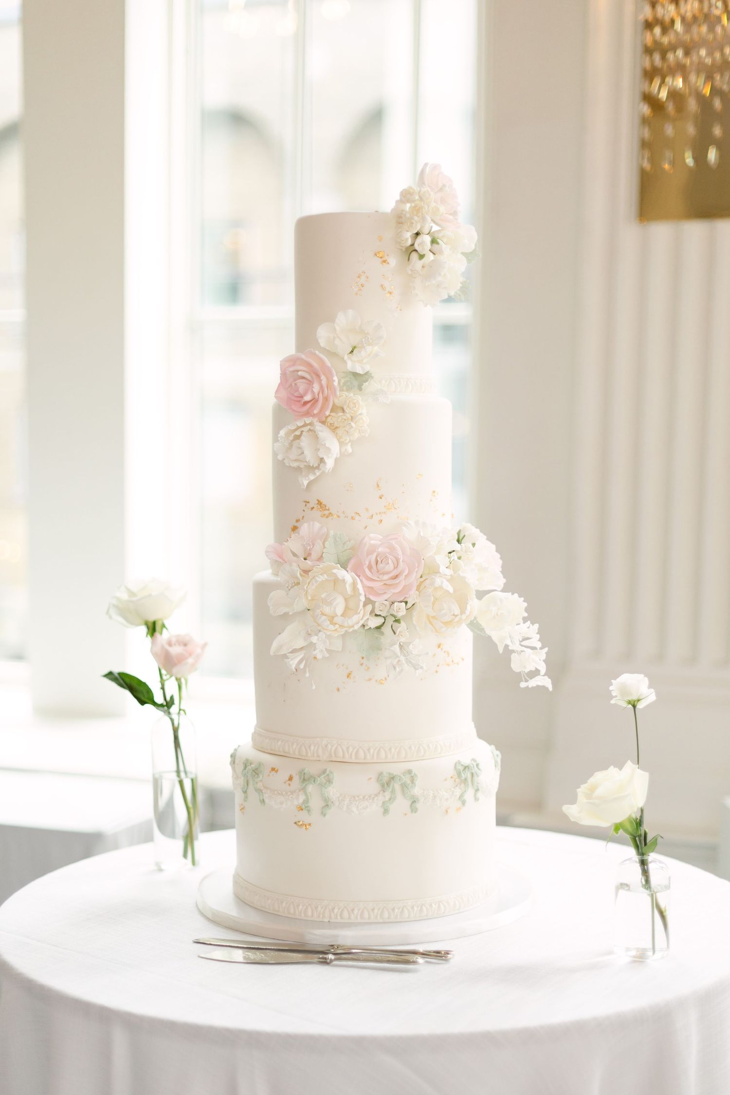 Elegant white wedding cake with delicate pink rose details displayed on a white pedestal against a bright window backdrop.