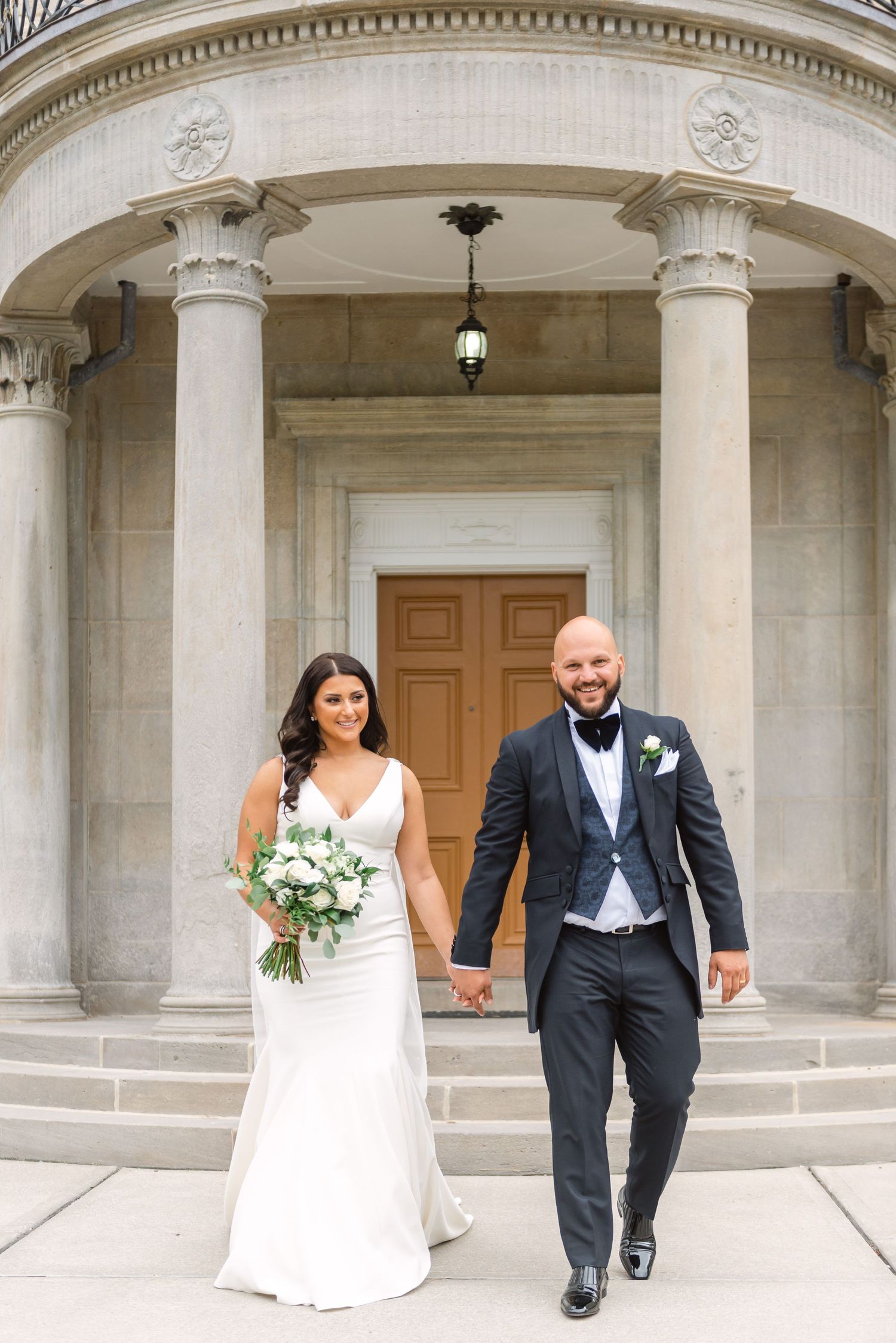 Wedding couple walking under classical stone colonnade with white pillars.