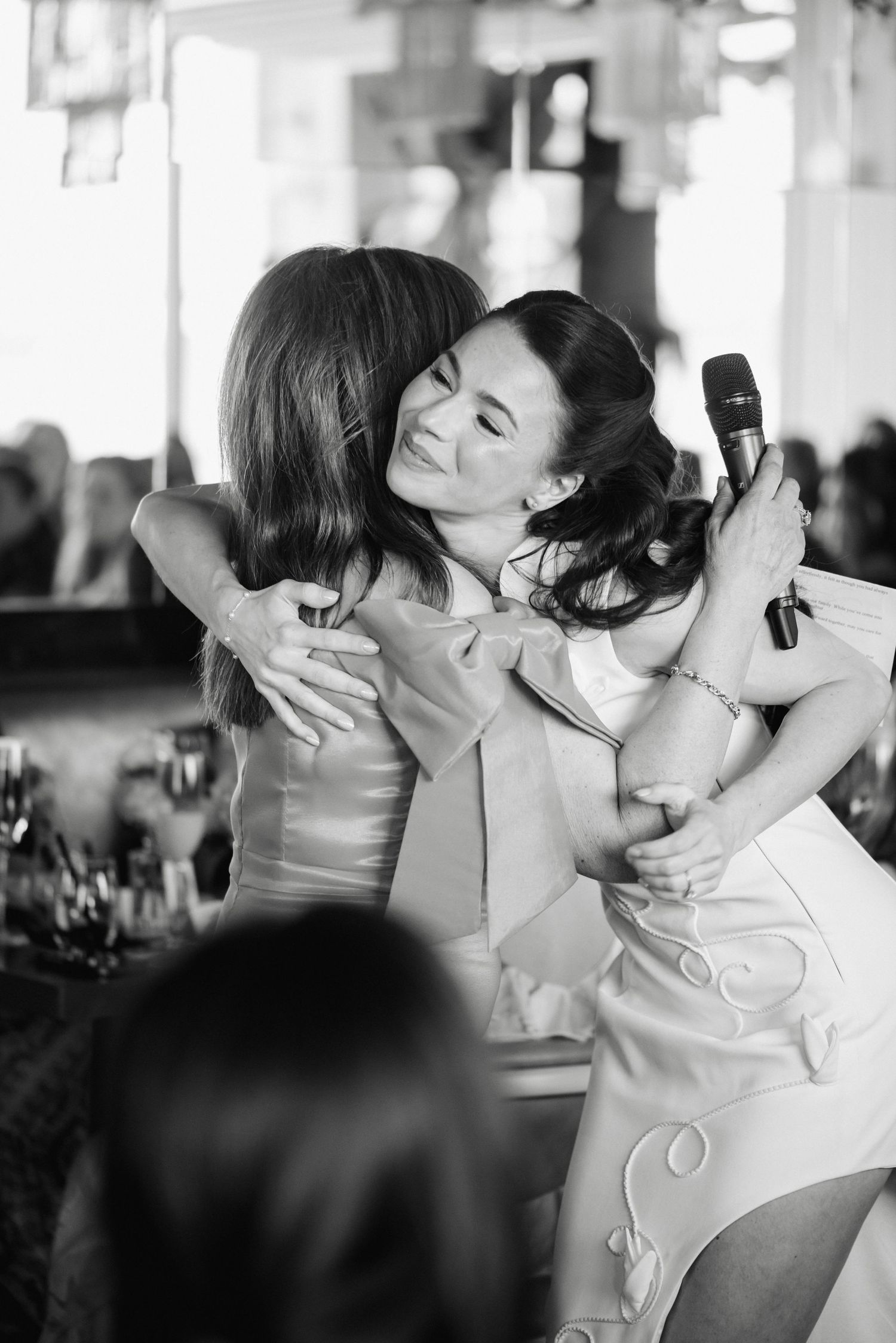 Two women share a warm embrace at a wedding reception, one holding a microphone during a heartfelt toast.