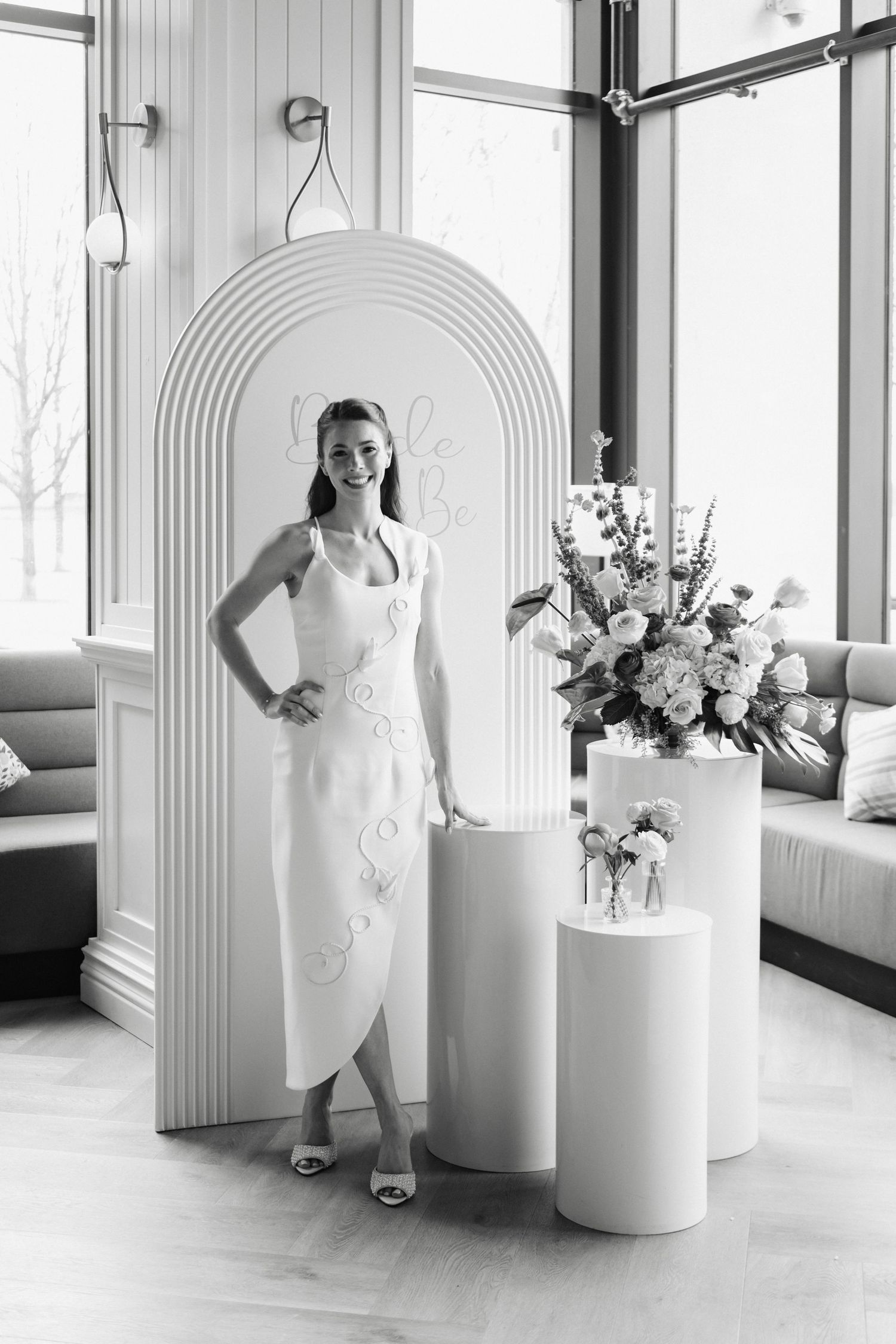 Woman in white midi dress smiling beside floral arrangement and arch backdrop in elegant black and white photo.