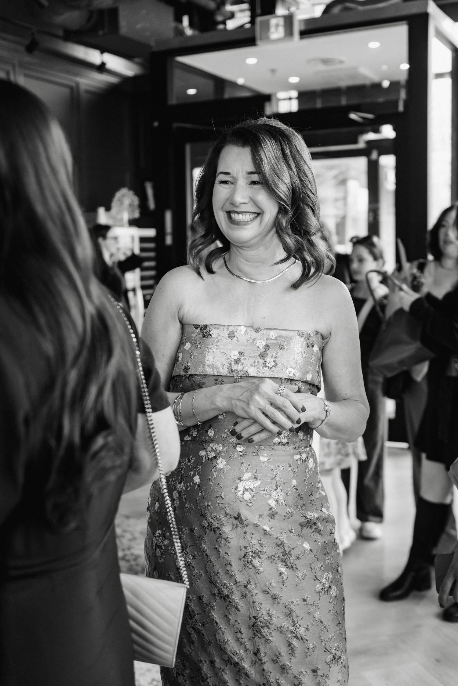 A smiling woman in a strapless floral dress at an elegant social event, wearing a necklace and ring.